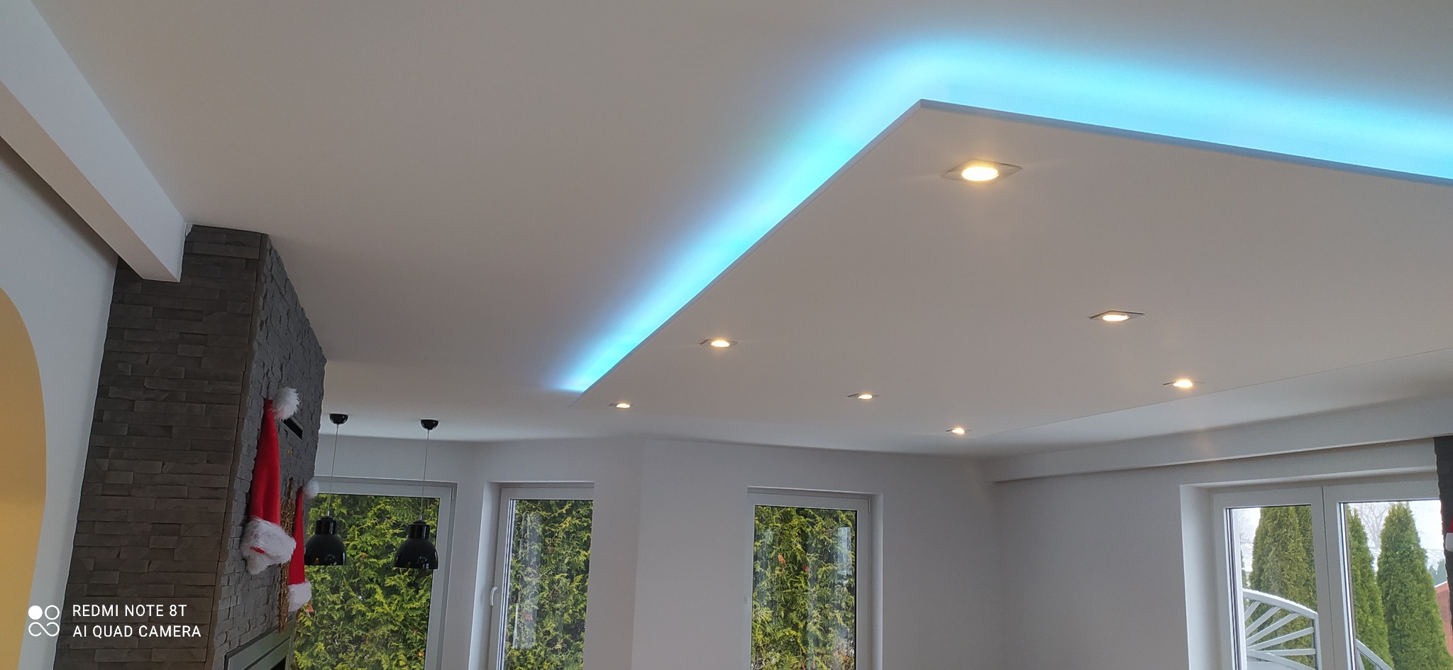 Nowoczesny sufit podwieszany z oświetleniem LED w kolorze niebieskim i punktowymi lampami halogenowymi, widoczne okna z zielenią za nimi i fragment ściany z kamienia dekoracyjnego z zawieszonymi...