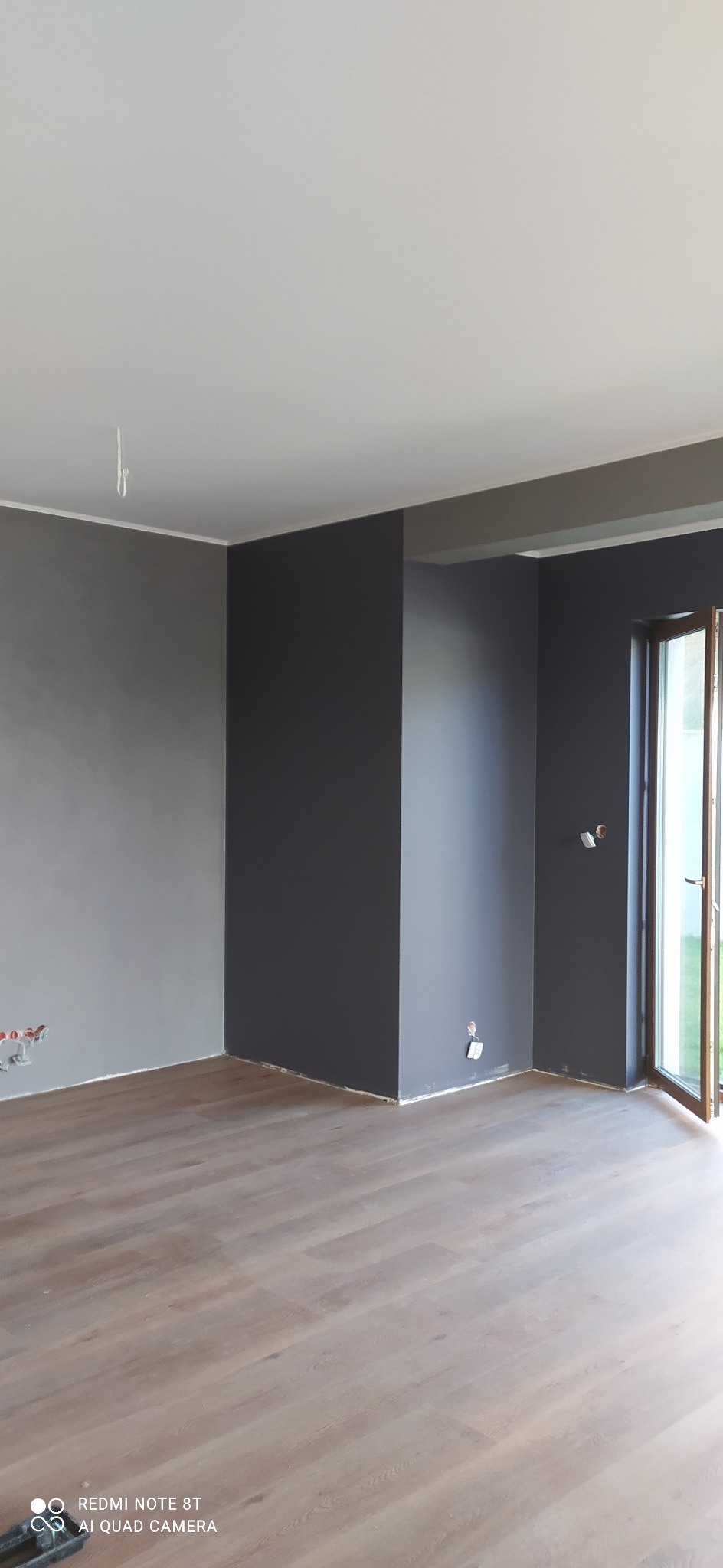 Wnętrze pokoju po malowaniu ścian na szaro i grafitowo, widoczne fragmenty instalacji elektrycznej, podłoga wyłożona panelami.