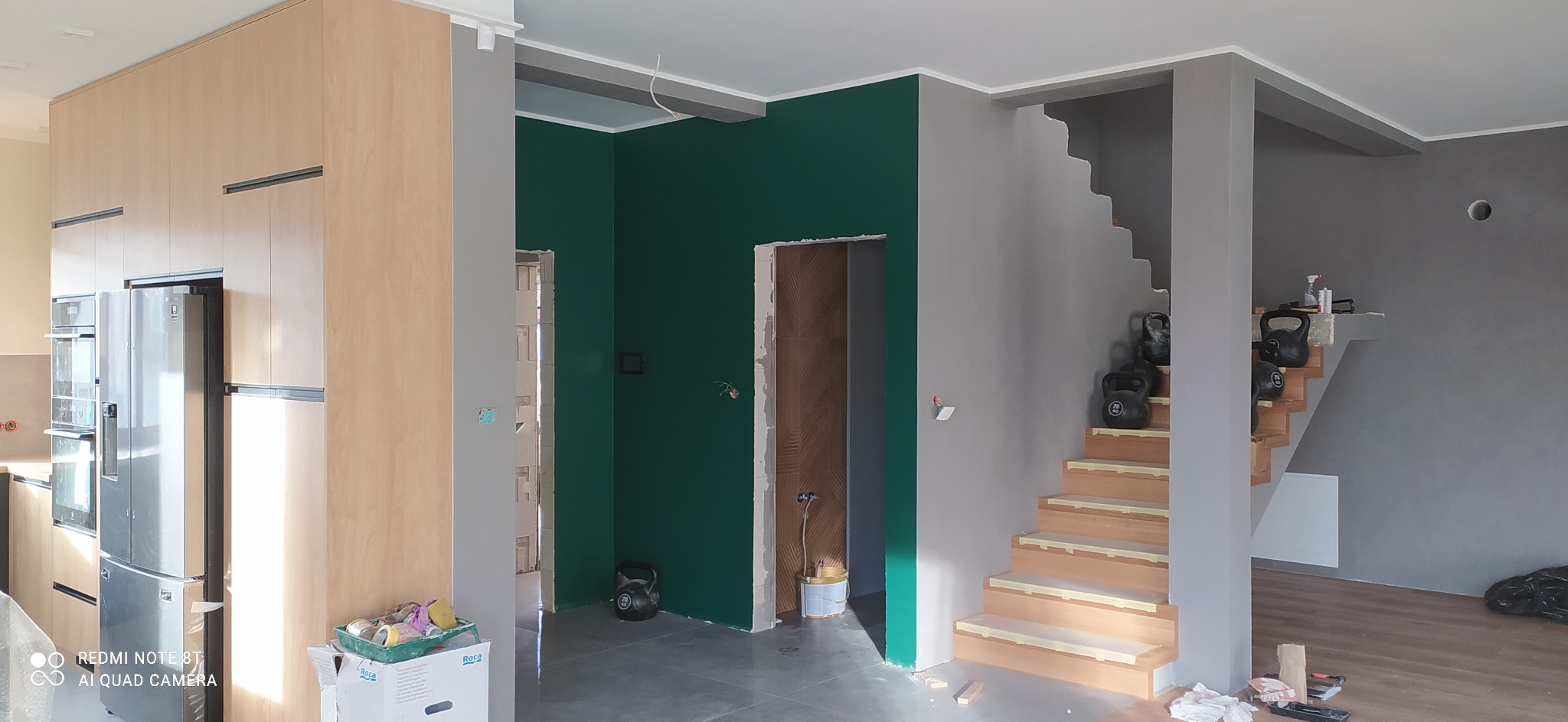 Wnętrze w trakcie remontu: pomalowana na szmaragdowo ściana, surowa klatka schodowa z obciążnikami, widoczna kuchnia z drewnianymi szafkami i srebrną lodówką, szary kolor ścian w tle.