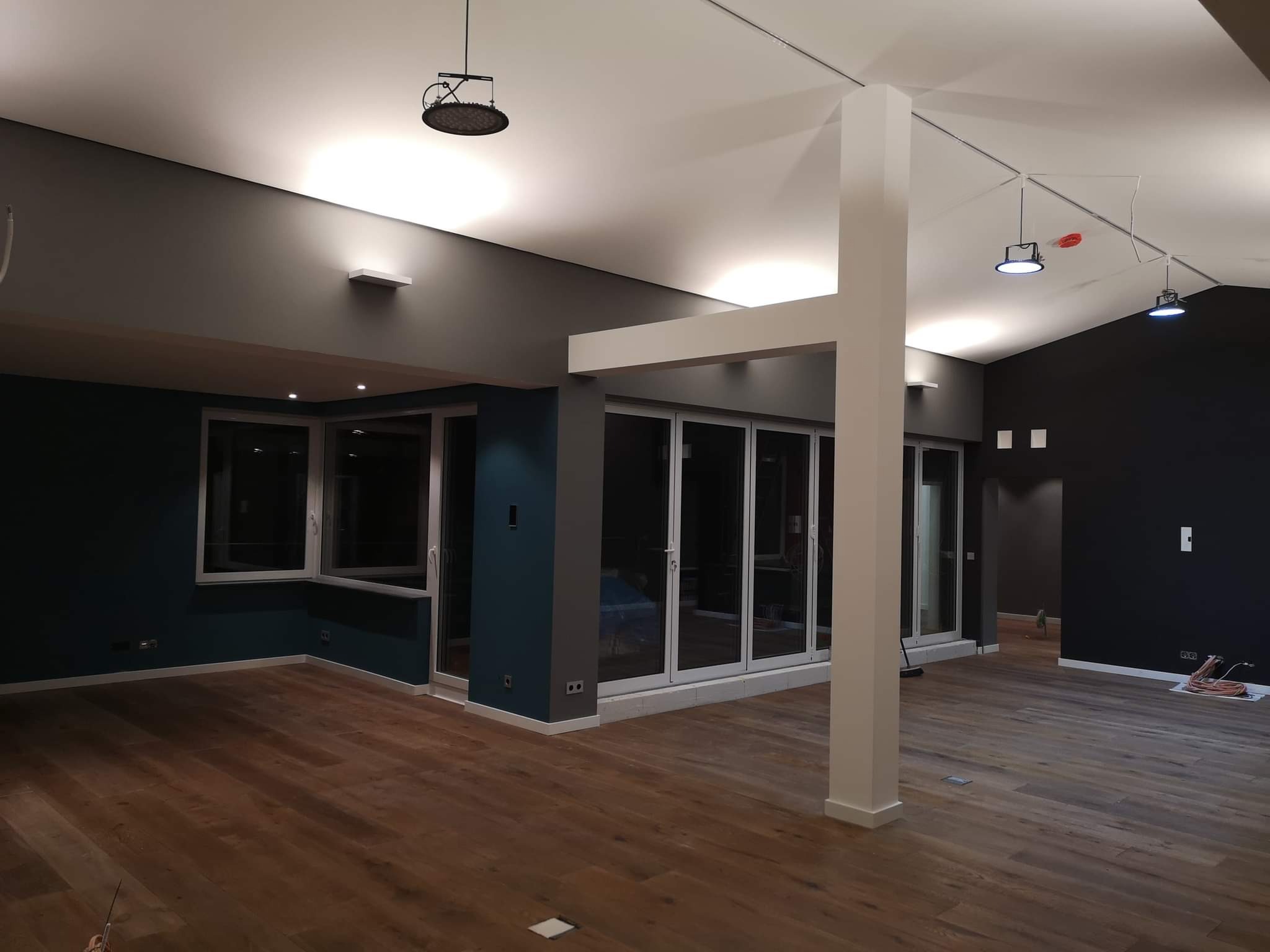 Wnętrze po remoncie z drewnianą podłogą, ciemnymi ścianami w odcieniach szarości i granatu, białymi oknami balkonowymi i minimalistycznym oświetleniem sufitowym.