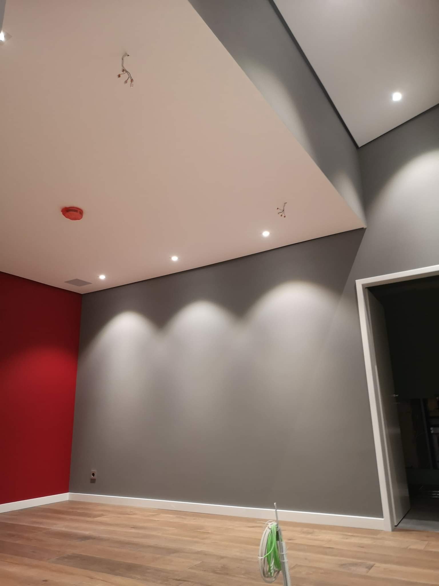 Wnętrze po remoncie: czerwona ściana kontrastująca z szarą, drewniana podłoga, biały sufit z punktowym oświetleniem i wystającymi przewodami, widoczne listwy przypodłogowe.