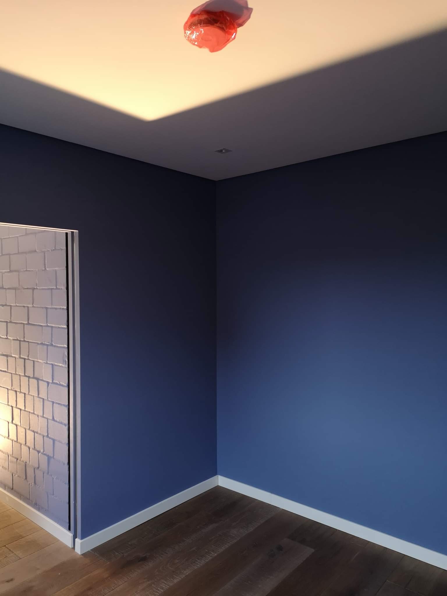 Pomieszczenie po malowaniu ścian na granatowo, zabezpieczona folią lampa sufitowa, widoczna imitacja cegieł na ścianie obok drzwi, drewniana podłoga.