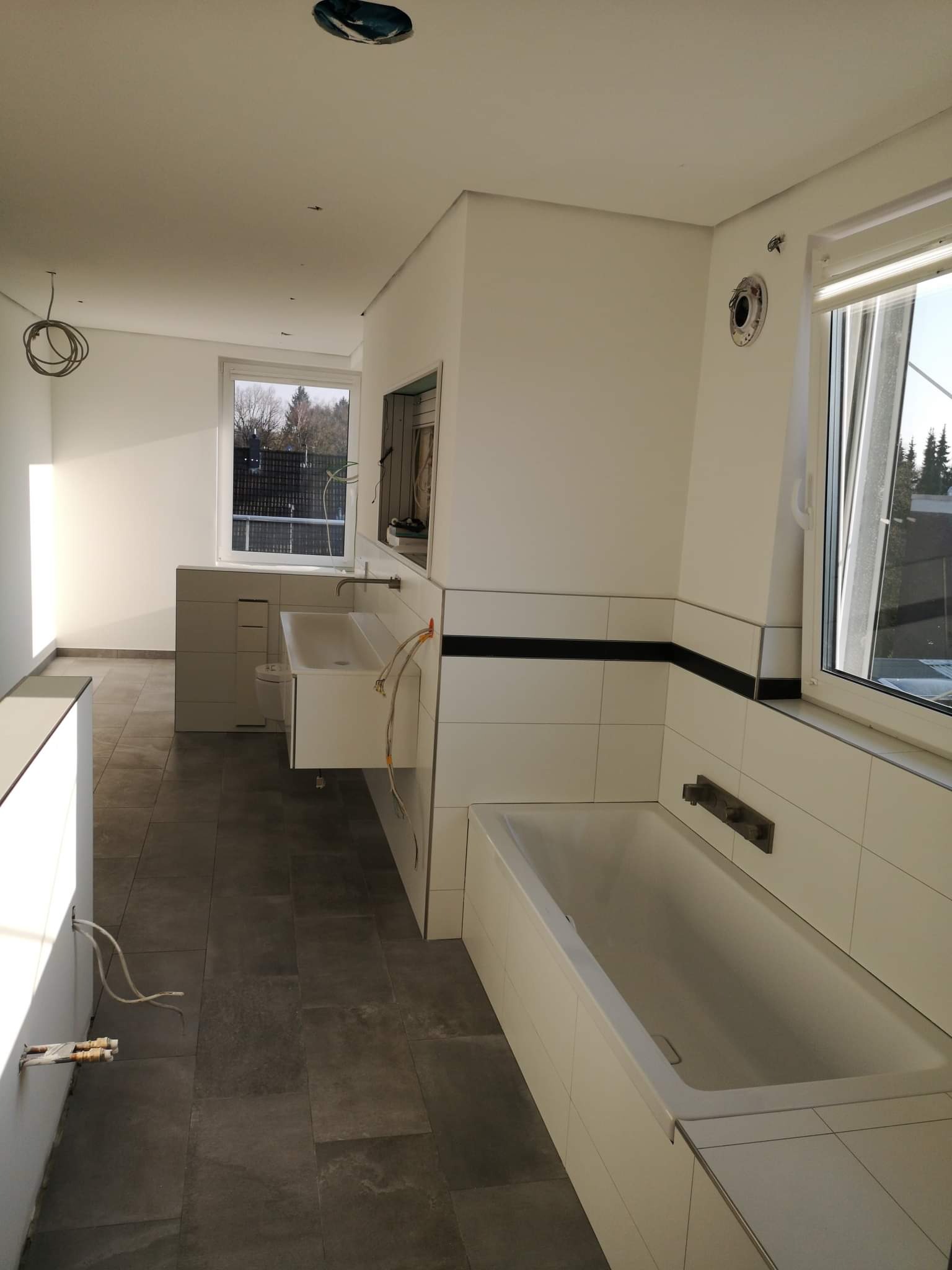 Wnętrze łazienki w trakcie wykańczania, widoczne białe płytki z czarnym pasem, wanna, umywalka z szafką, szare płytki podłogowe, wystające kable i otwory w ścianach i suficie czekające na osprzęt.