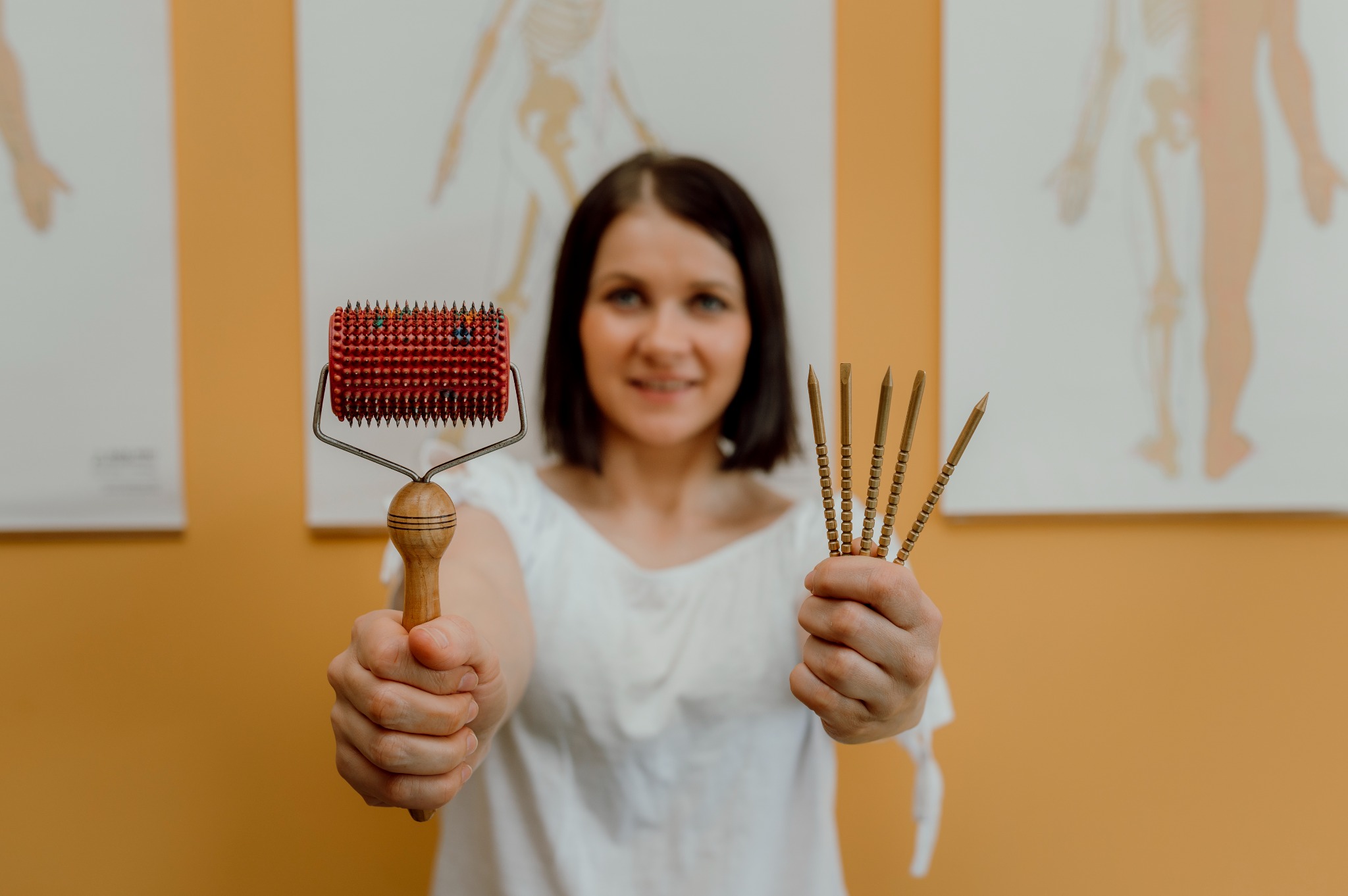 Kobieta prezentuje narzędzia do akupunktury: czerwony wałek z kolcami i zestaw złotych igieł na tle schematycznych rysunków szkieletu.