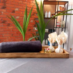 Przygotowany zestaw do zabiegu: zwinięty ciemny ręcznik, dwa bawełniane stemple ziołowe i złoty świecznik na drewnianej tacy, na tle ceglanej ściany i półki z kosmetykami.