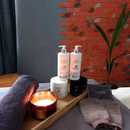 A&P Atelier Beauty Massage - Przygotowane stanowisko do zabiegów pielęgnacyjnych: kosmetyki APIS, aromatyczna świeca, rękawice do peelingu, ręcznik i voucher na masaż, ułożone na drewnianej tacy na tle ceglanej ściany...