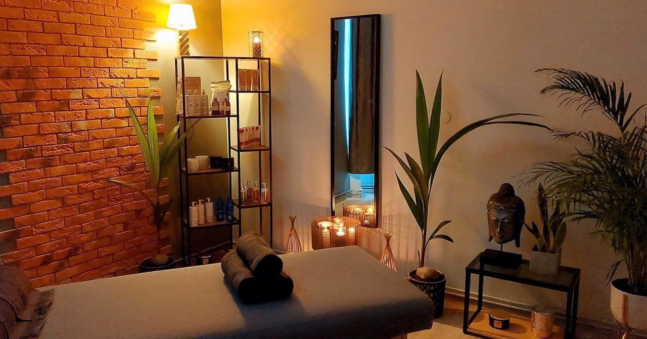 Przytulne wnętrze gabinetu z łóżkiem do masażu, ceglaną ścianą i dekoracjami w stylu orientalnym, w tle regał z kosmetykami i podświetlane lustro.