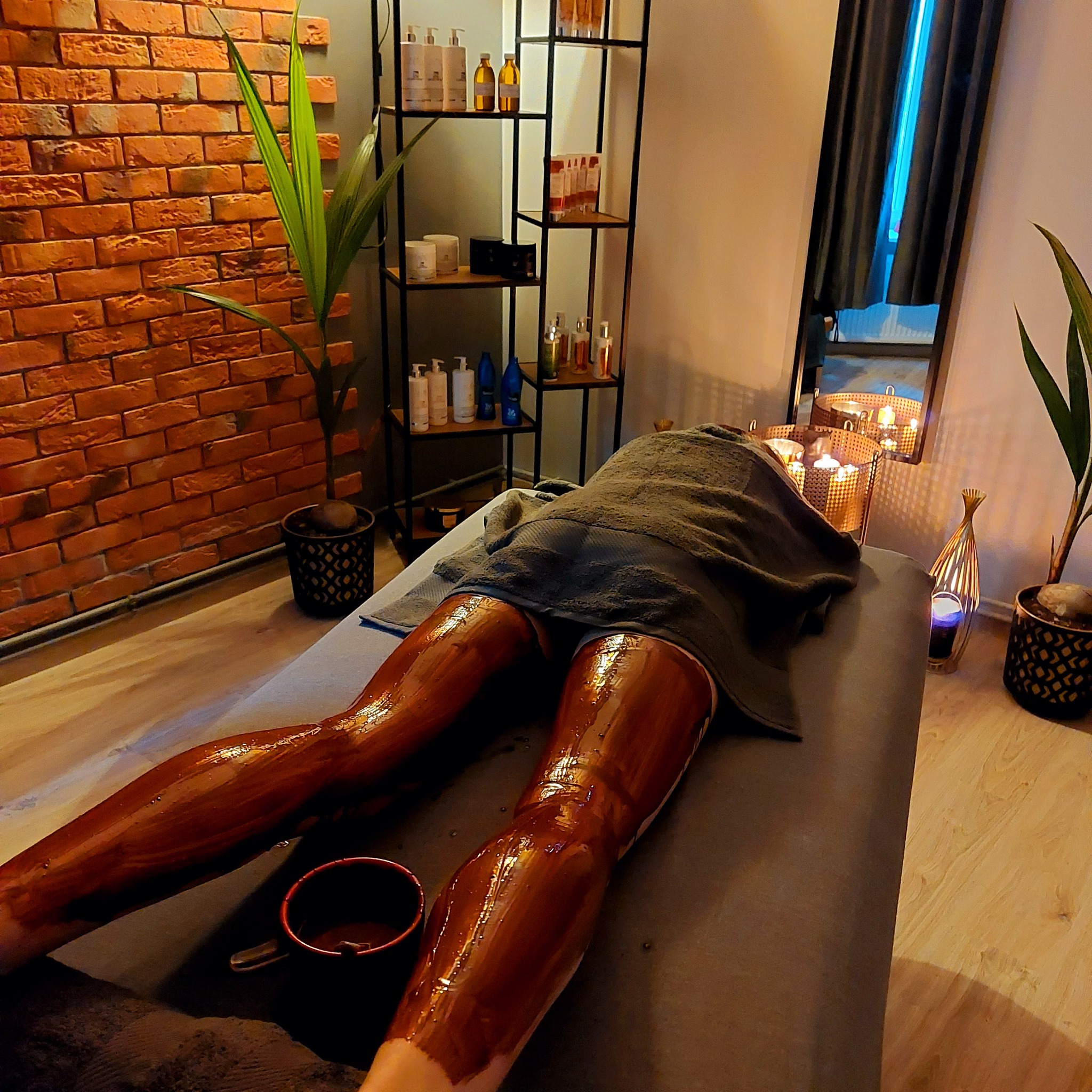 Klientka leżąca na stole do zabiegów, z nogami pokrytymi czekoladową maską, w tle ceglana ściana i dekoracyjny regał z kosmetykami.