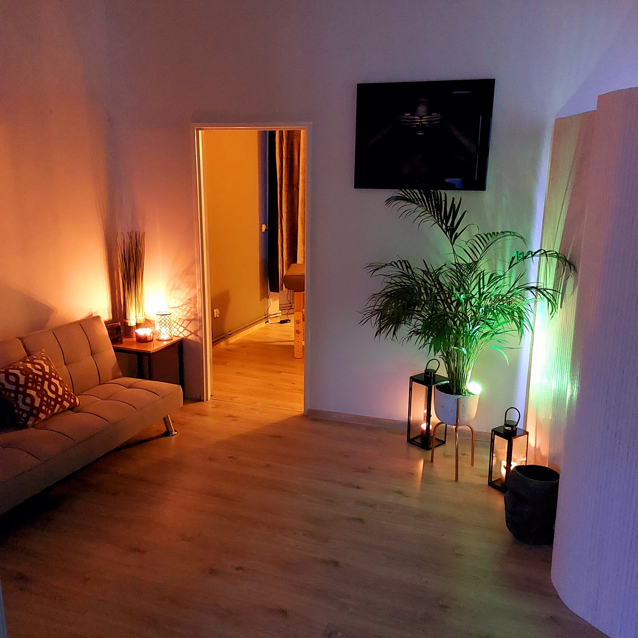 Przytulne wnętrze gabinetu z sofą, stolikiem z dekoracjami, rośliną w doniczce i świecami, w tle widoczny stół do masażu.