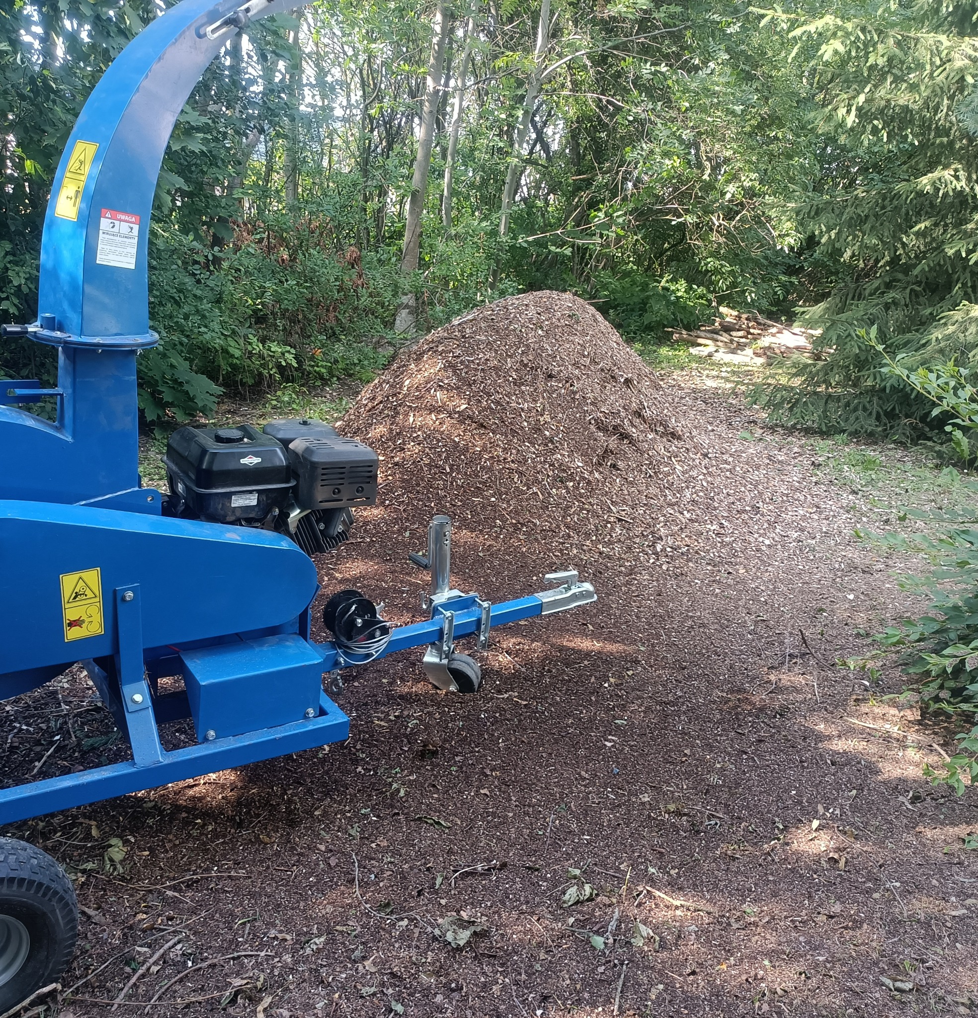 Niebieski rębak do gałęzi stoi obok dużej pryzmy zrębków drzewnych na leśnej ścieżce.