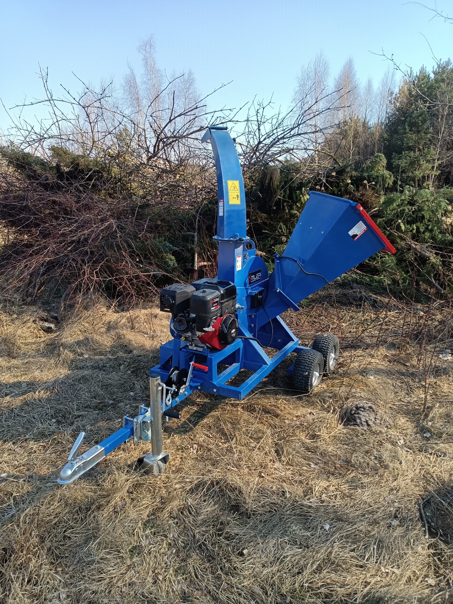 Niebieska, mobilna rębarka do gałęzi marki TLEA z silnikiem spalinowym, zaparkowana na trawniku obok sterty gałęzi, gotowa do pracy.
