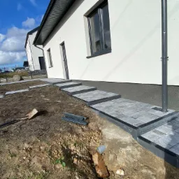 Schody wejściowe do domu wyłożone szarą kostką brukową, widoczne narzędzia i niedokończone prace ziemne w otoczeniu budynku.