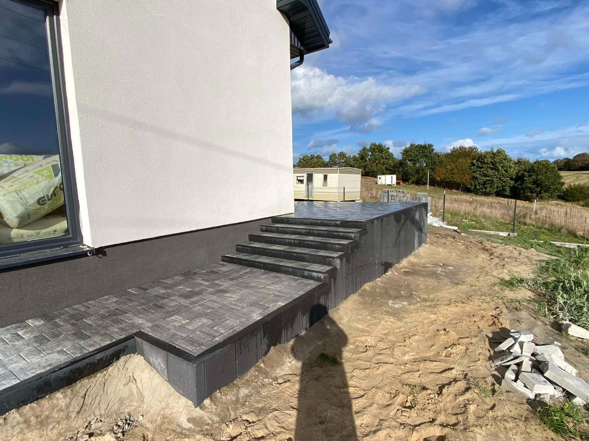 Wykończone schody i taras z ciemnoszarej kostki brukowej przy elewacji domu, widoczne worki z materiałami budowlanymi i teren w trakcie prac ziemnych.