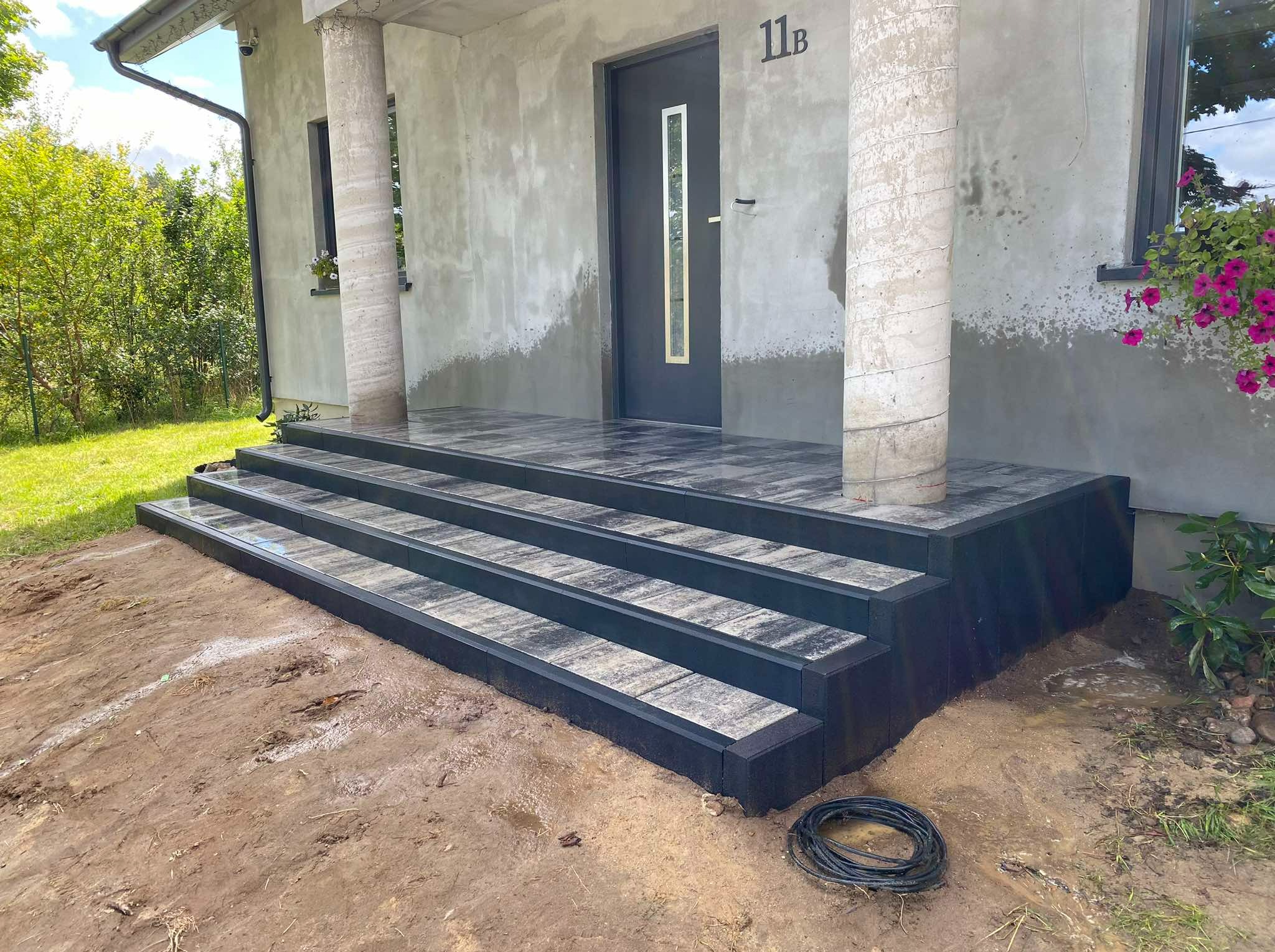 Schody wejściowe do domu wyłożone szarą kostką z czarnymi obrzeżami, widoczne ślady prac ziemnych i zwój kabla na pierwszym planie.