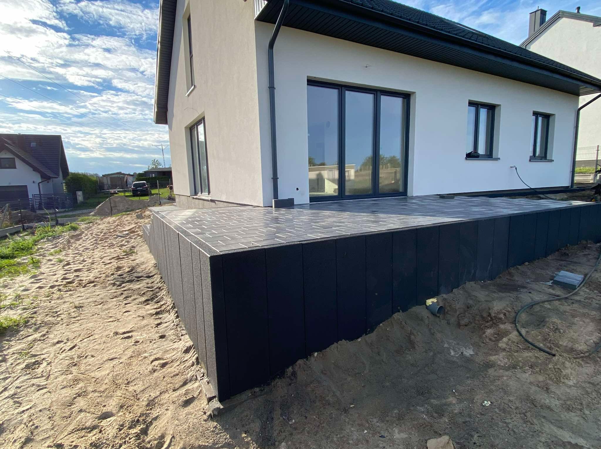 Nowoczesny taras z szarej kostki brukowej przy białym domu z czarnymi ramami okiennymi, widoczne niedokończone prace ziemne wokół tarasu.