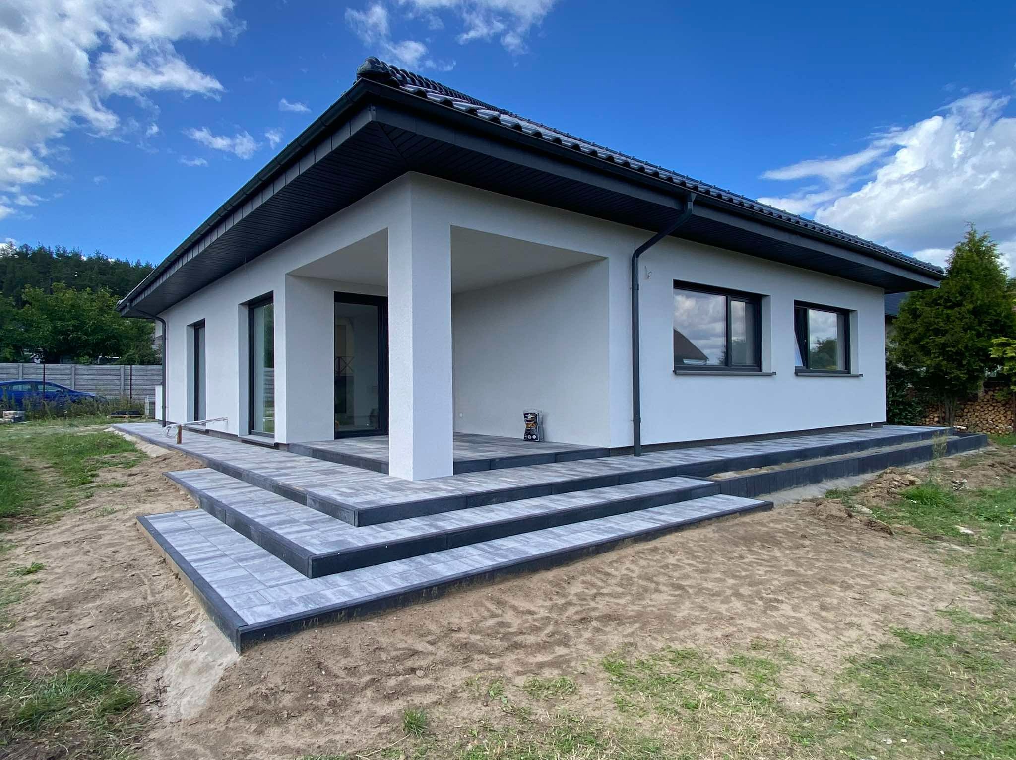 Nowoczesny dom jednorodzinny z tarasem i schodami wyłożonymi szarą kostką brukową z ciemnymi obrzeżami, widok z poziomu gruntu na tle błękitnego nieba.