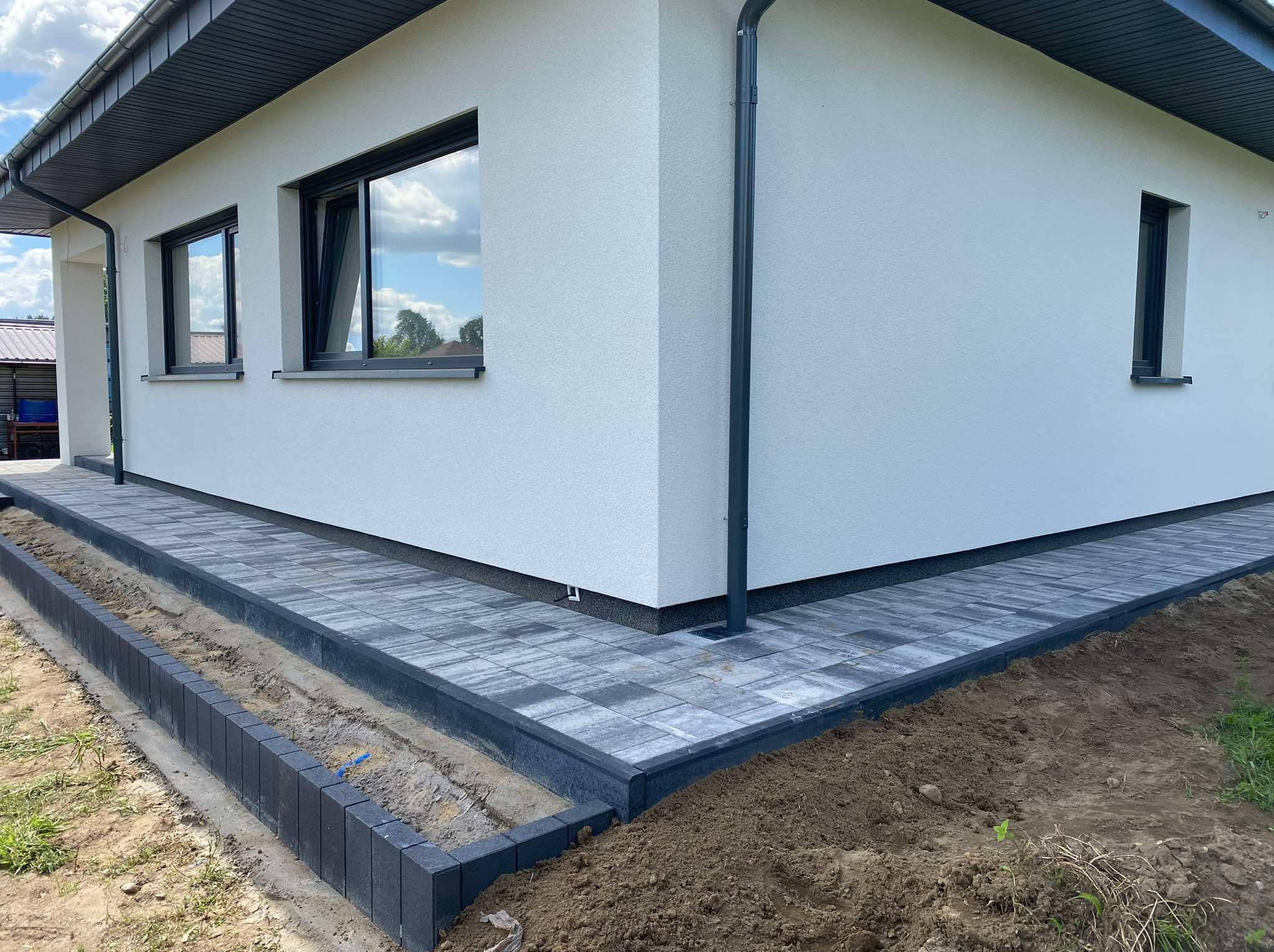 Szara kostka brukowa ułożona wokół narożnika białego domu z czarnymi oknami i rynnami, widoczny fragment rabaty z obrzeżem z kostki.