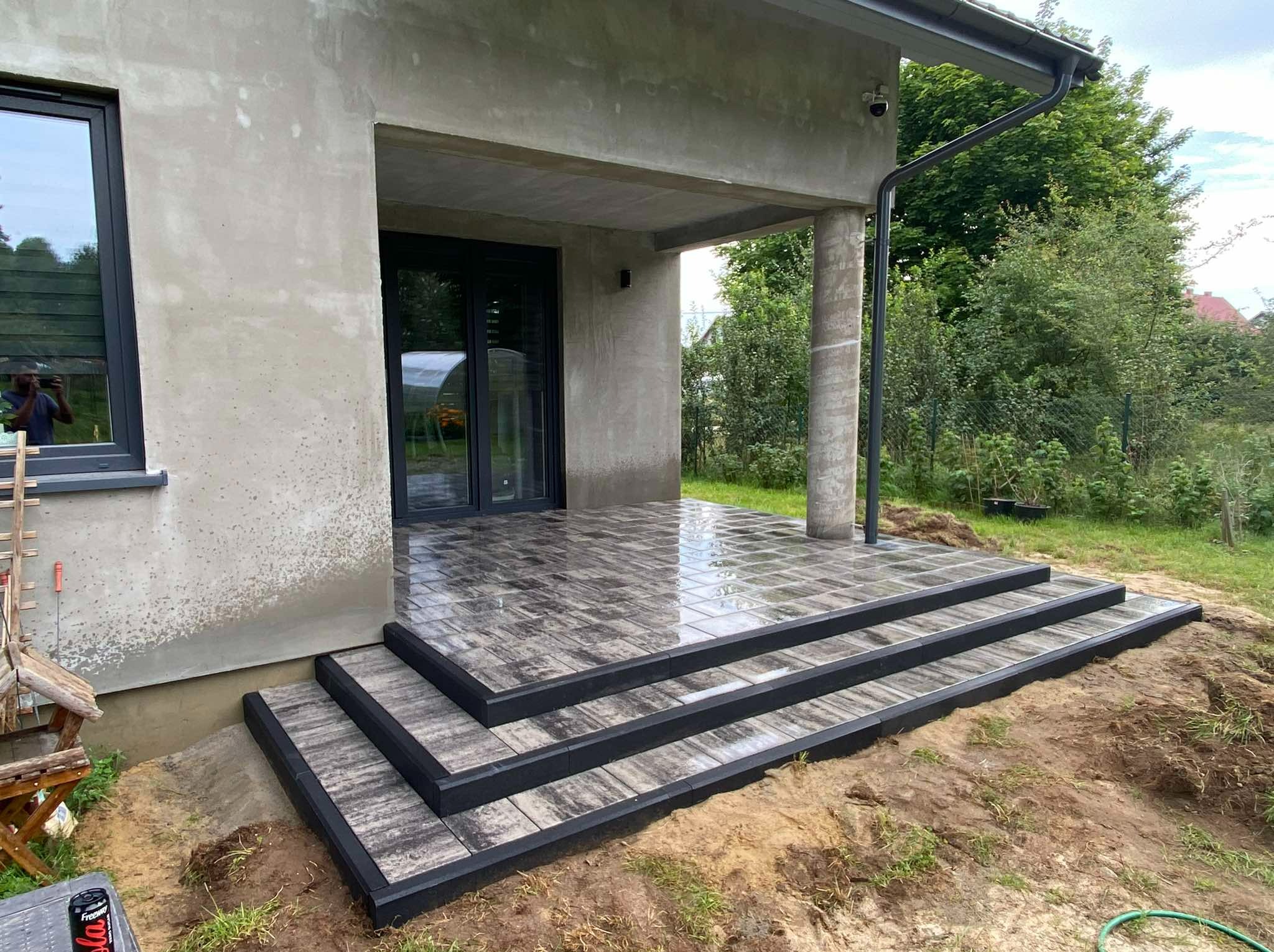 Schody wejściowe wyłożone szarą kostką brukową z czarnym obramowaniem, prowadzące do domu z szarą elewacją i czarnymi oknami, widok z boku na tle zieleni.