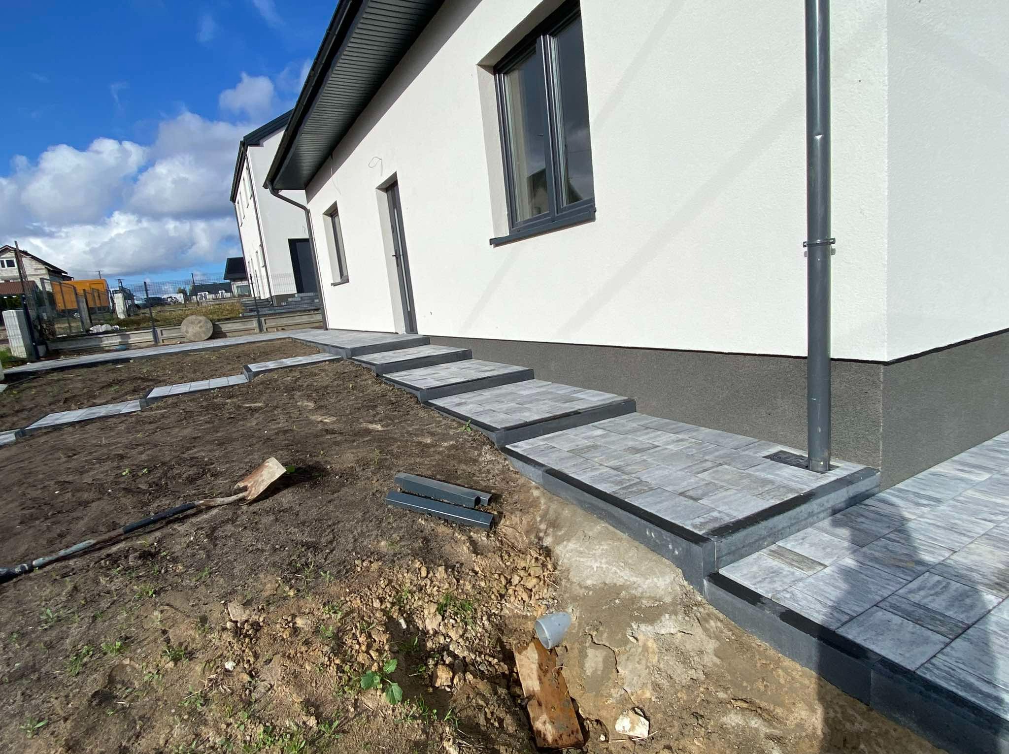 Schody wejściowe do domu wyłożone szarą kostką brukową, widoczne narzędzia i niedokończone prace ziemne w otoczeniu budynku.
