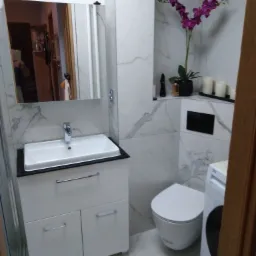 Nowoczesna łazienka z marmurowymi płytkami, umywalką na szafce z białymi frontami, toaletą i pralką, ozdobiona fioletową orchideą i świecami.