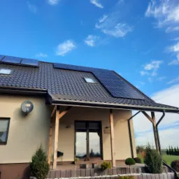 Panele słoneczne zamontowane na dachu domu z ciemnej dachówki, widok z dołu na tle błękitnego nieba z delikatnymi obłokami, antena satelitarna na ścianie budynku.