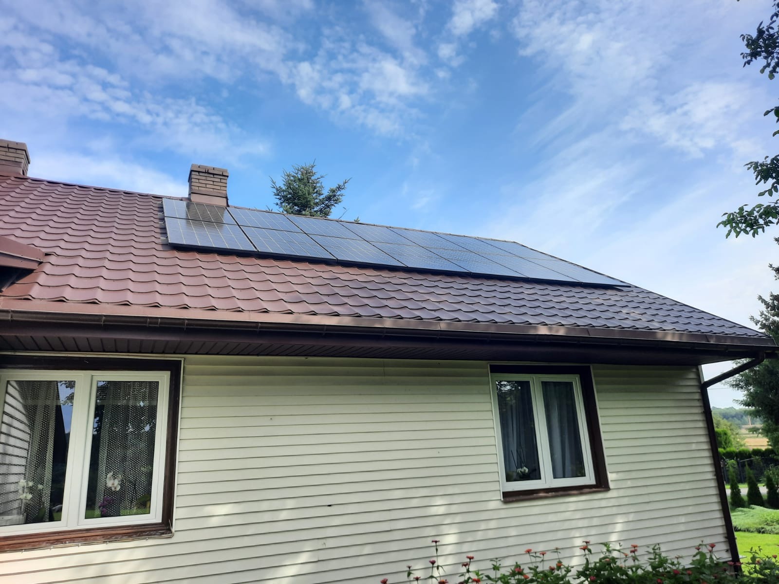 Panele słoneczne zamontowane na brązowym dachu domu jednorodzinnego z widocznymi dwoma kominami i białymi oknami, na tle błękitnego nieba z chmurami.