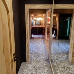 Remys - Otwarta drewniana rama drzwi z dekoracyjnym wzorem,  ukazująca wzorzystą podłogę z płytek w głębi wnętrza. Lustro po prawej stronie odbija widok.