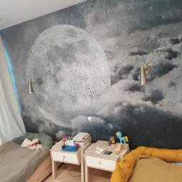 Pokój dziecięcy z fototapetą przedstawiającą księżyc i chmury, oświetlony dwoma kinkietami w kolorze mosiądzu, z dwoma łóżkami i szafkami nocnymi.