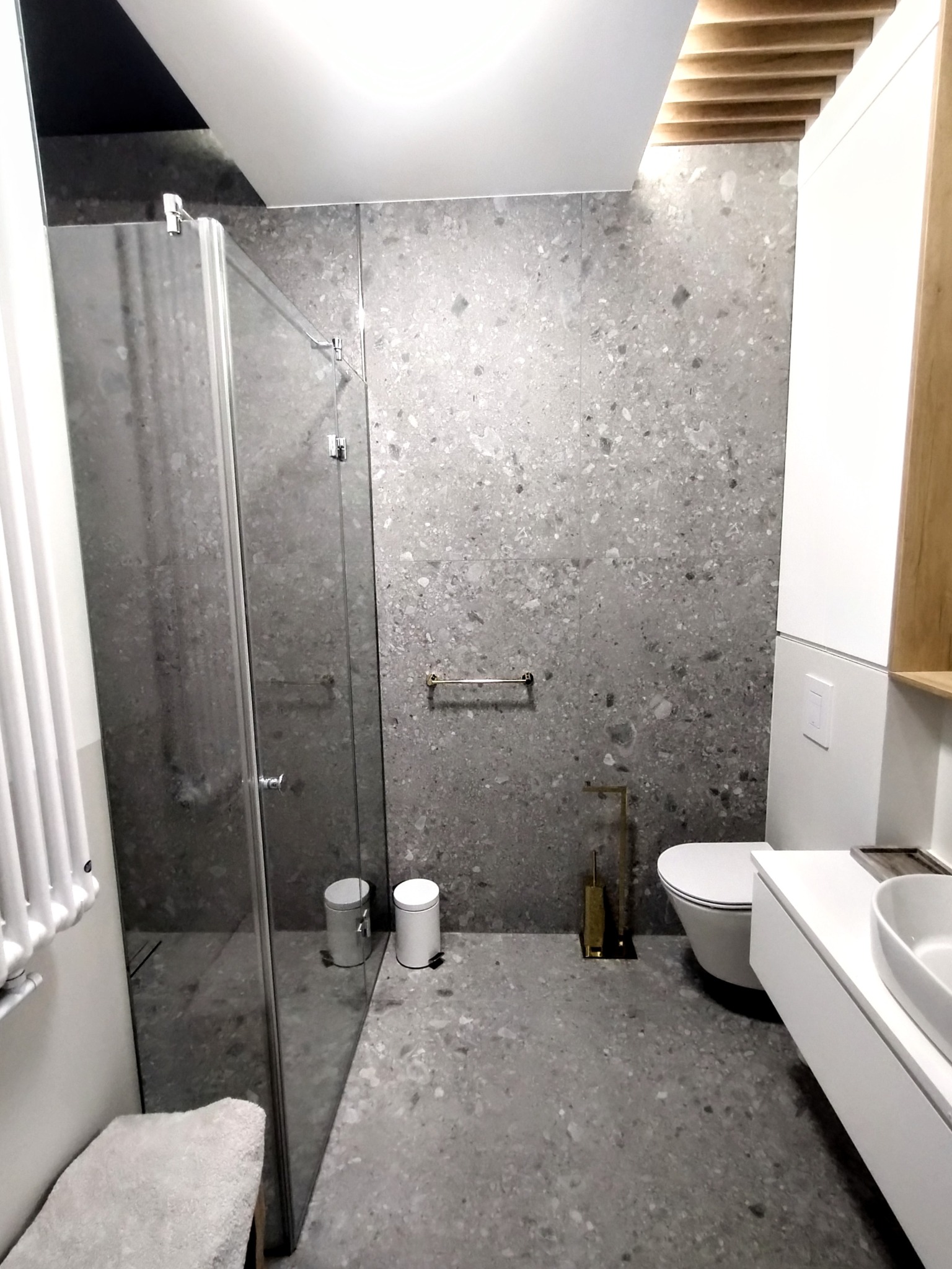 Nowoczesna łazienka z szarymi płytkami lastryko na ścianach i podłodze. Kabina prysznicowa ze szklanymi drzwiami, biała umywalka z szafką i toaleta.