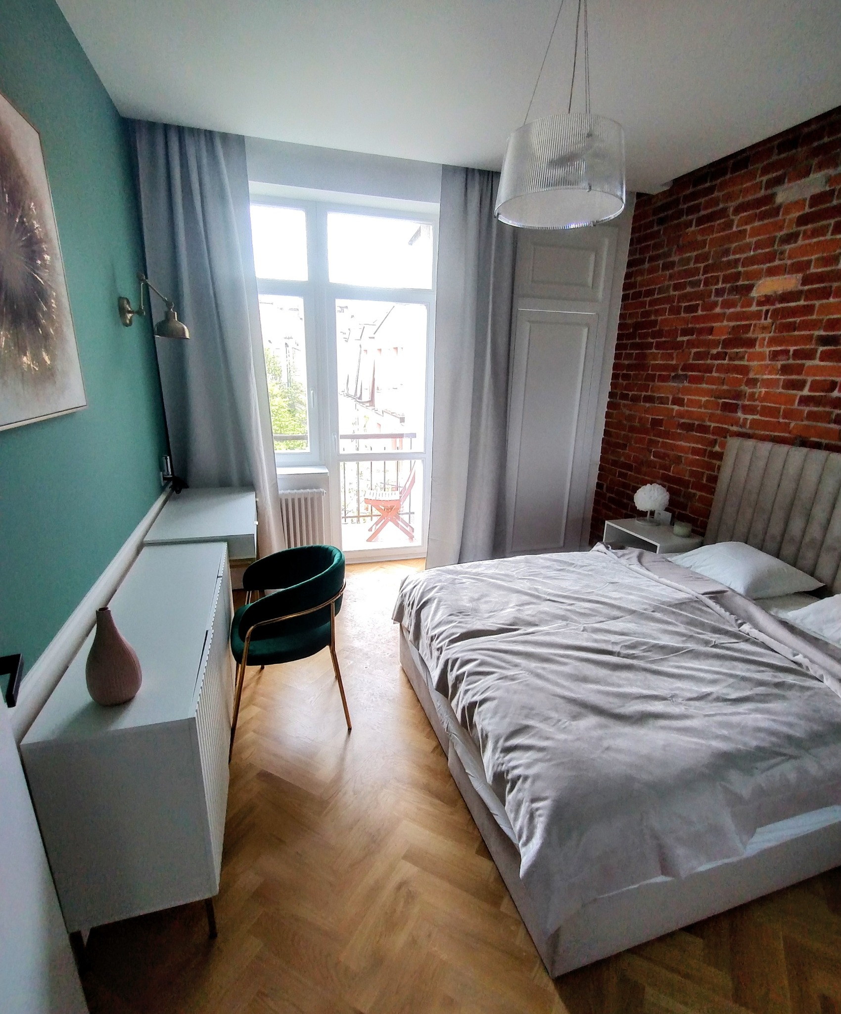 Sypialnia w stylu eklektycznym: zielona ściana, ceglana ściana, parkiet w jodełkę i balkon z widokiem na miasto. Krzesło z zielonego weluru.