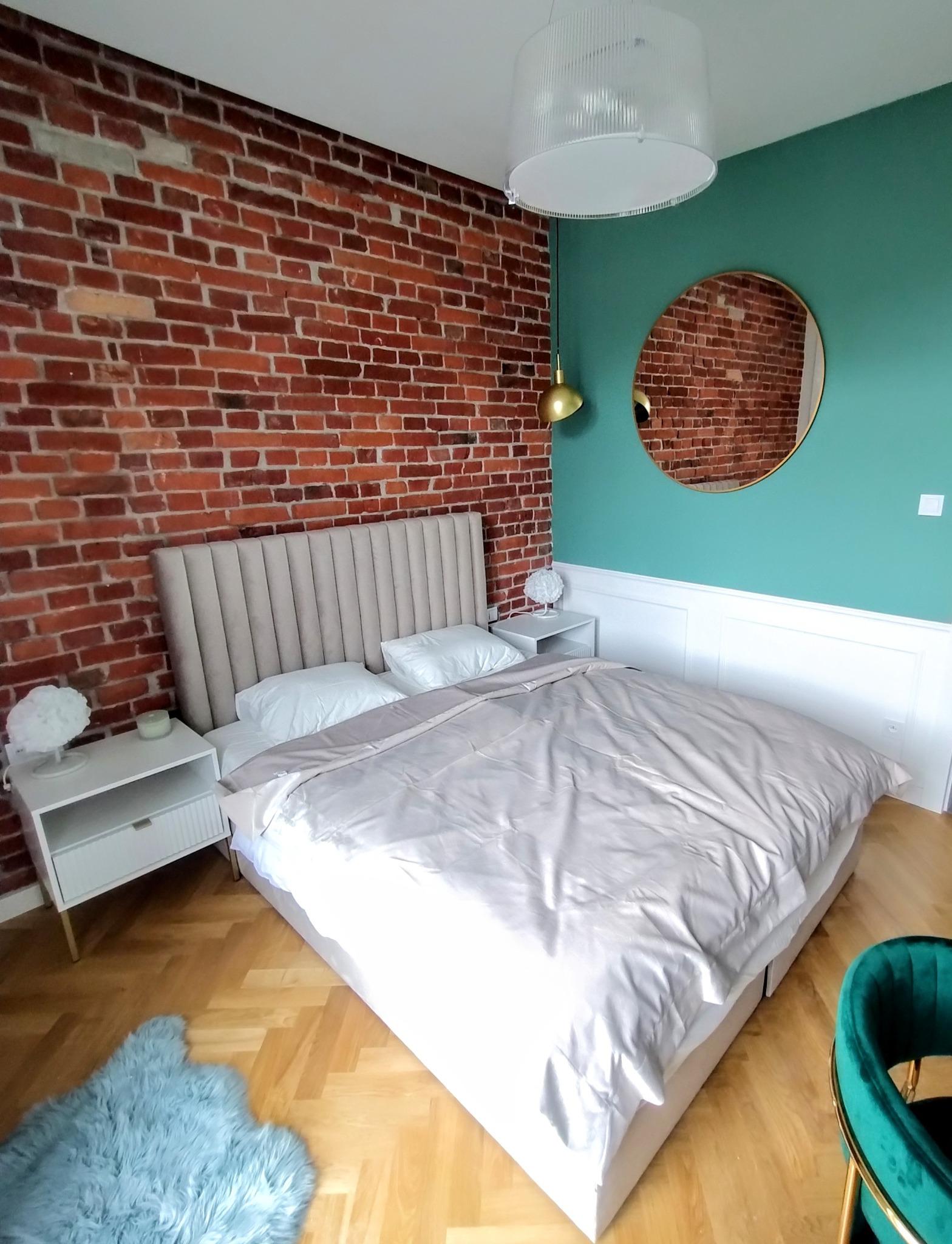 Sypialnia z ceglaną ścianą, łóżkiem z beżowym zagłówkiem i okrągłym lustrem na zielonej ścianie. Parkiet na podłodze i designerskie lampki nocne.