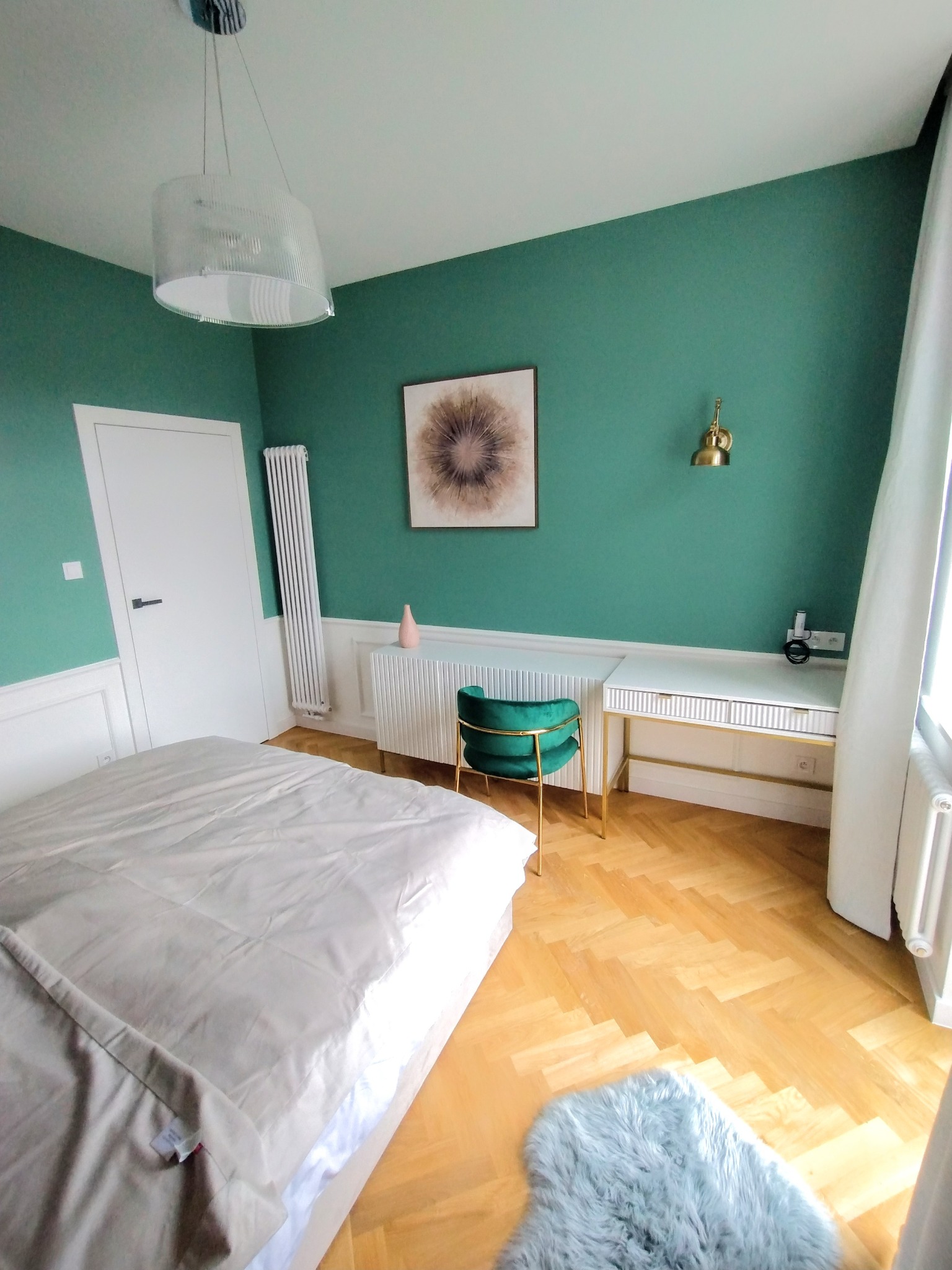 Jasne wnętrze sypialni w stylu retro: zielone ściany, parkiet w jodełkę i białe meble. Widoczne łóżko, biurko, krzesło i dekoracje ścienne. Całość w dziennym świetle.