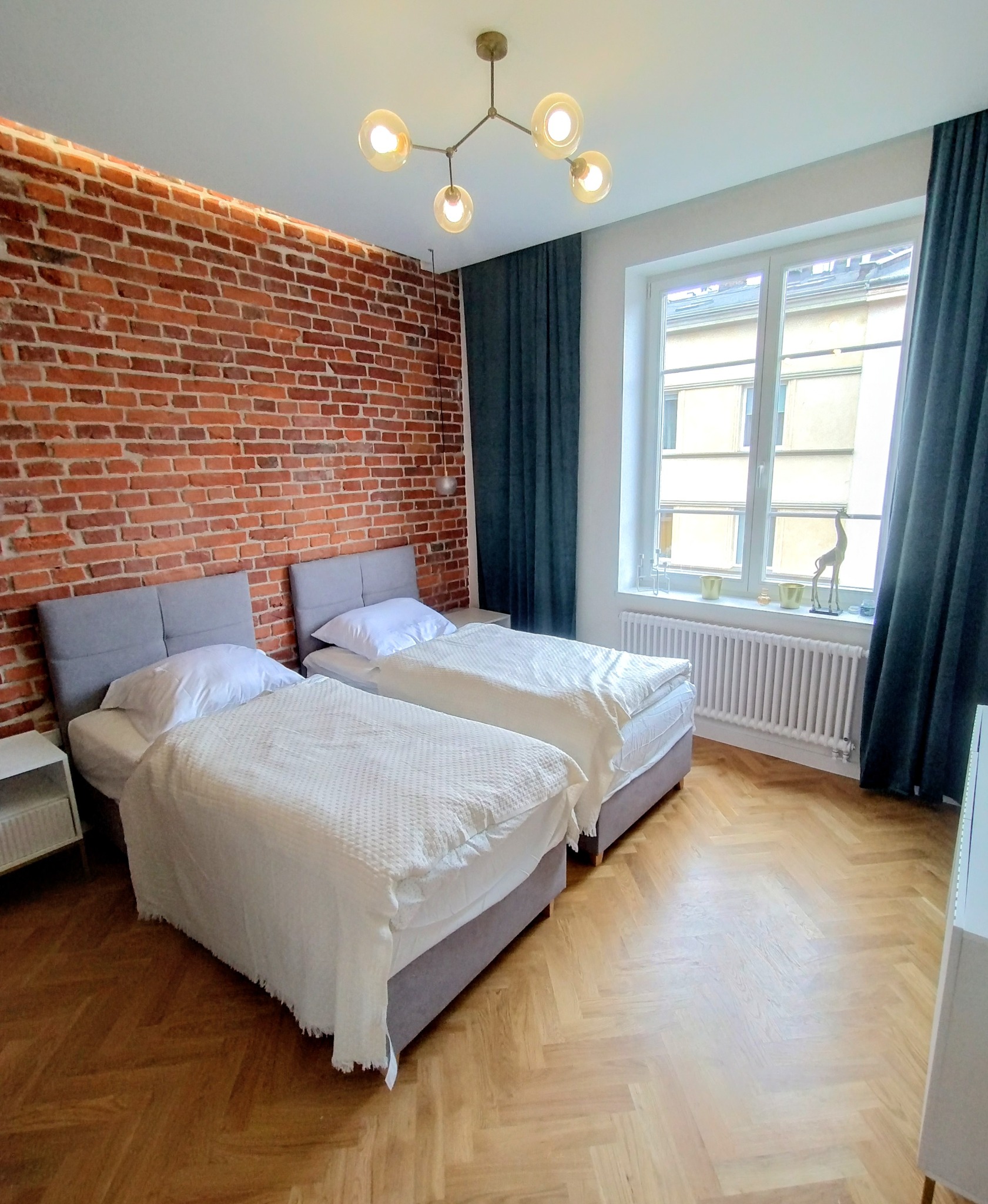 Pokój z dwoma łóżkami, ceglaną ścianą i parkietem w jodełkę. Nowoczesna lampa sufitowa i zasłony. Widok na budynek z okna.