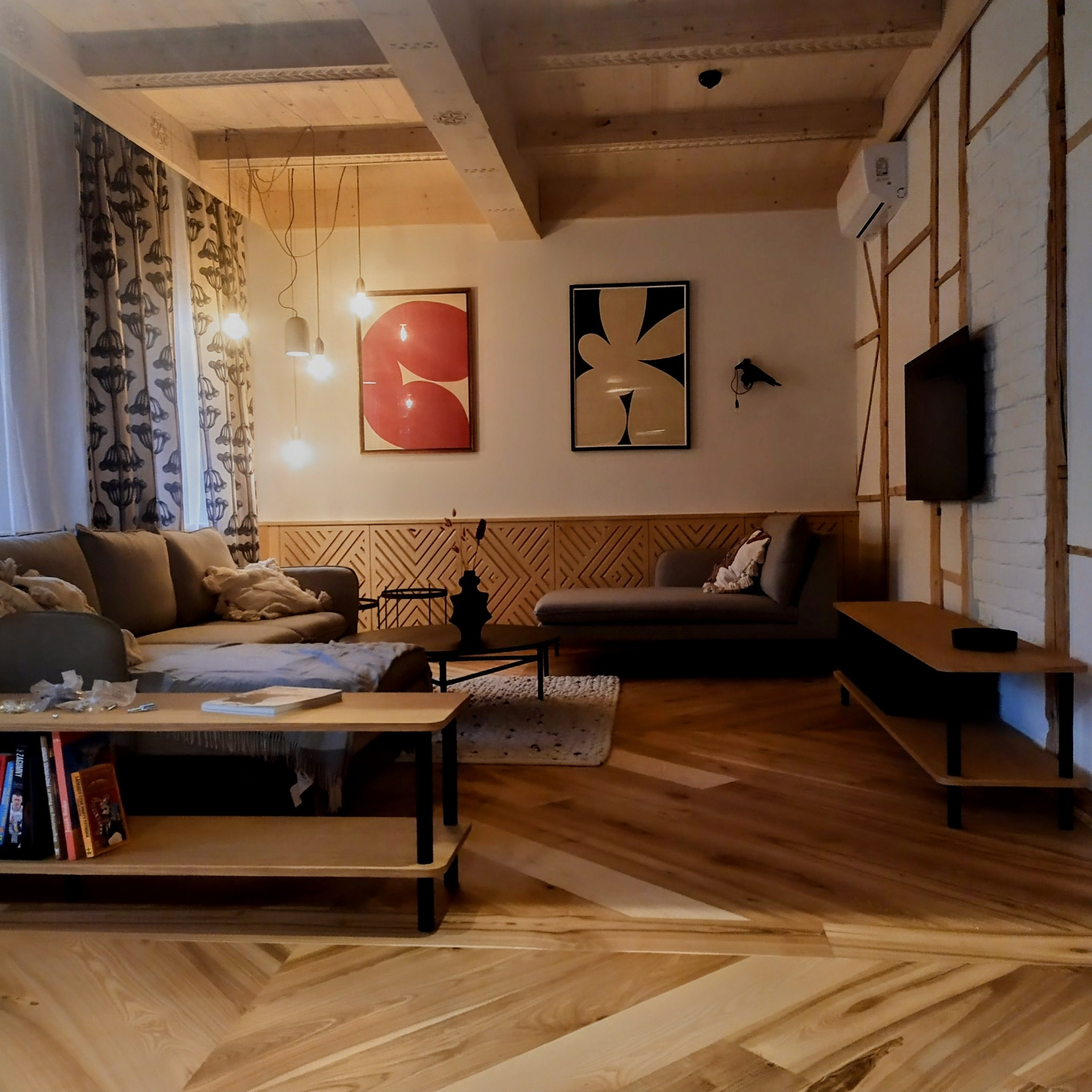 Stylowe wykończenie wnętrza z drewnianą podłogą w jodełkę, ozdobnymi panelami ściennymi, nowoczesnymi obrazami i klimatyzacją. Widok na salon z sofą i stolikami.