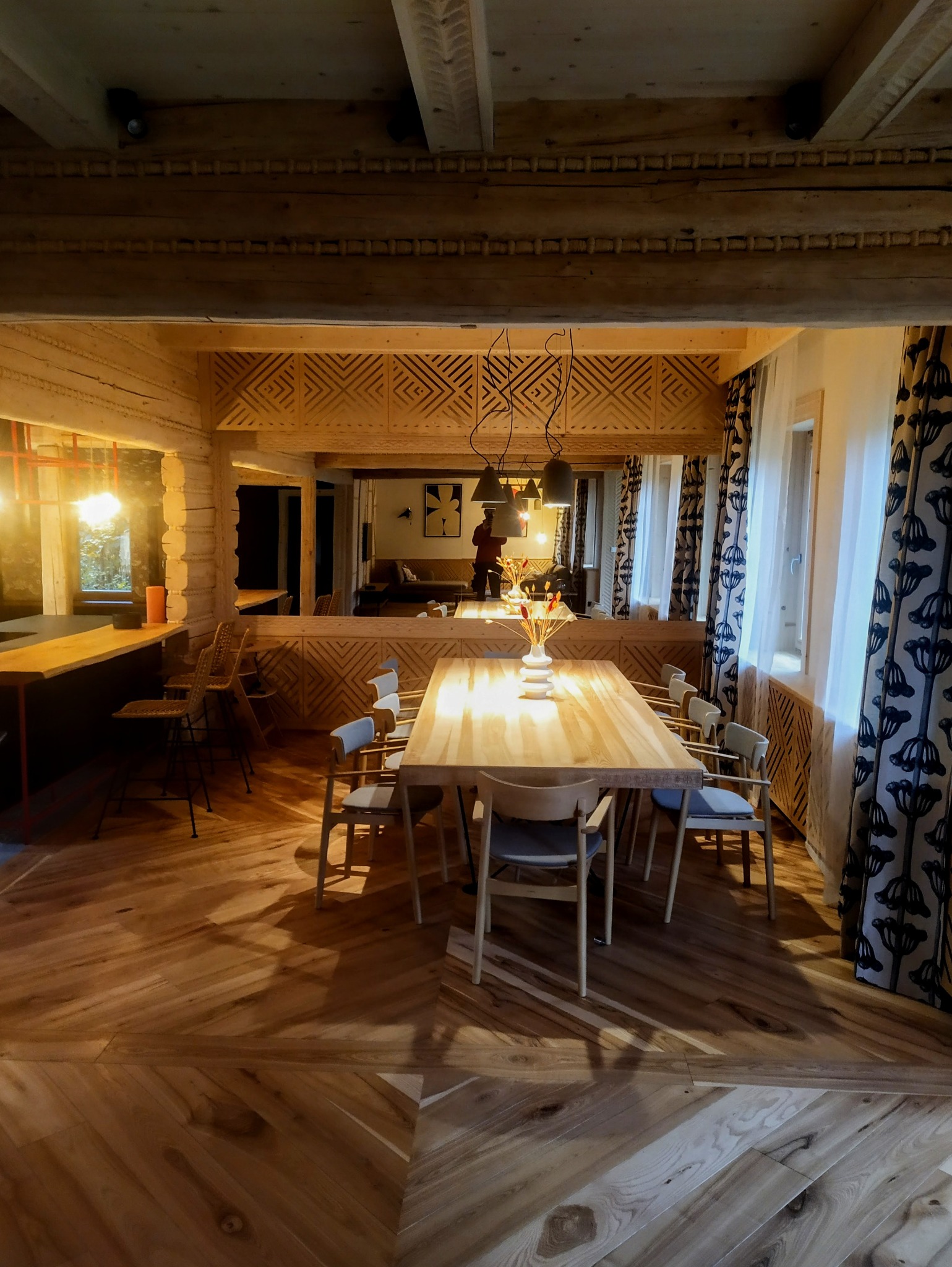 Drewniane wykończenie wnętrza jadalni z parkietem ułożonym we wzór chevron, drewnianym stołem i krzesłami. Dekoracyjne panele z geometrycznym wzorem.