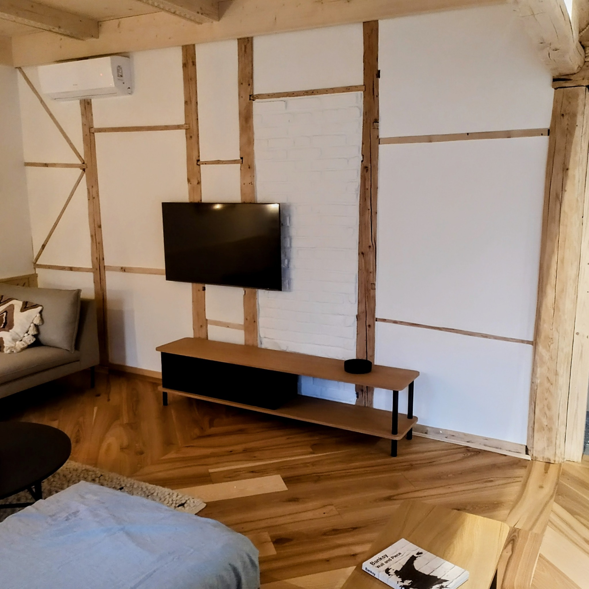 Wykończone wnętrze pokoju z drewnianą konstrukcją ścian, pomalowaną na biało cegłą i parkietem ułożonym w jodełkę. Telewizor na ścianie i meble w stylu skandynawskim.