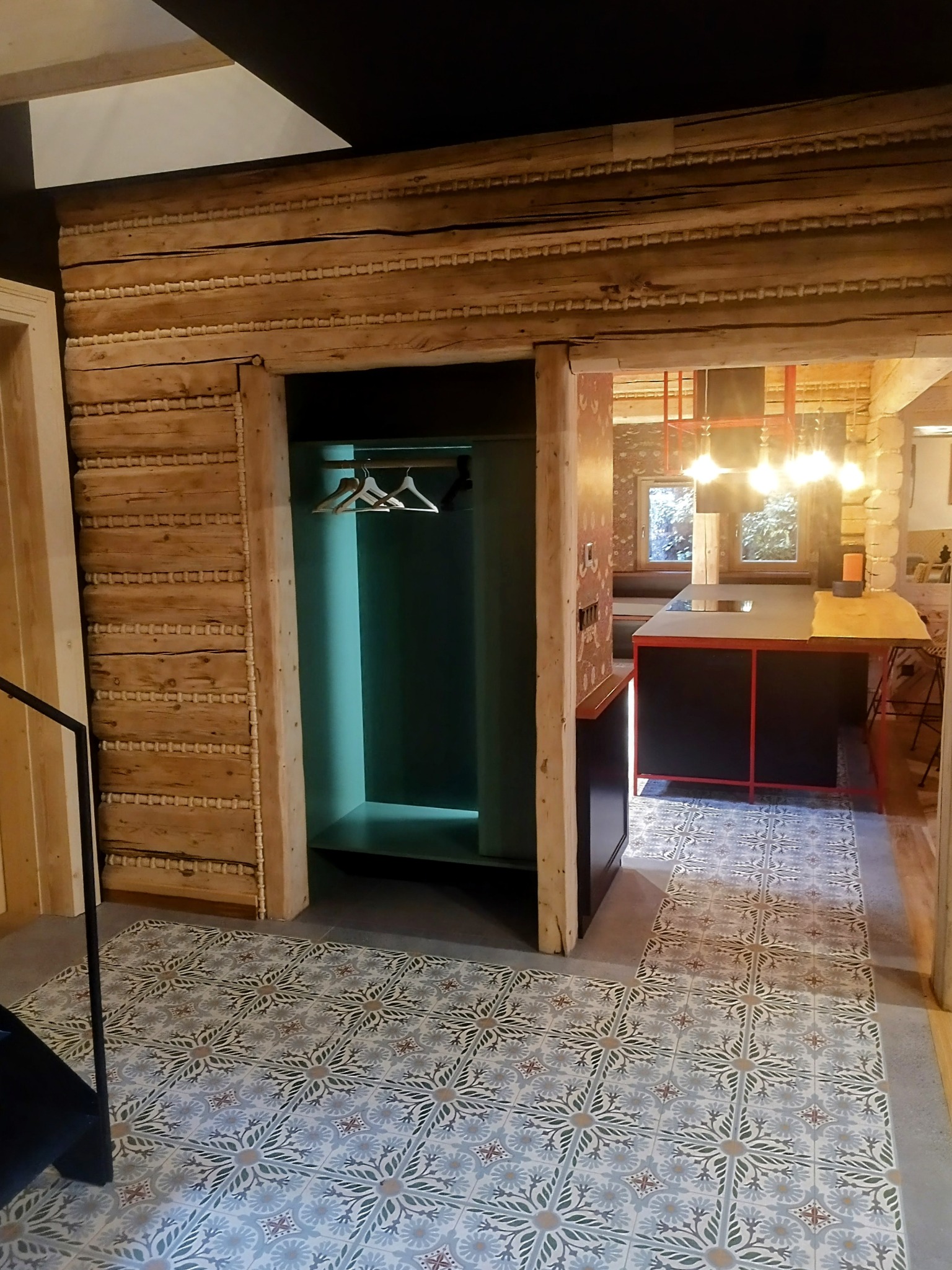 Wnętrze domu w stylu góralskim: drewniane ściany, wzorzysta podłoga z płytek, zabudowana szafa wnękowa z wieszakami i widok na kuchnię z wyspą.