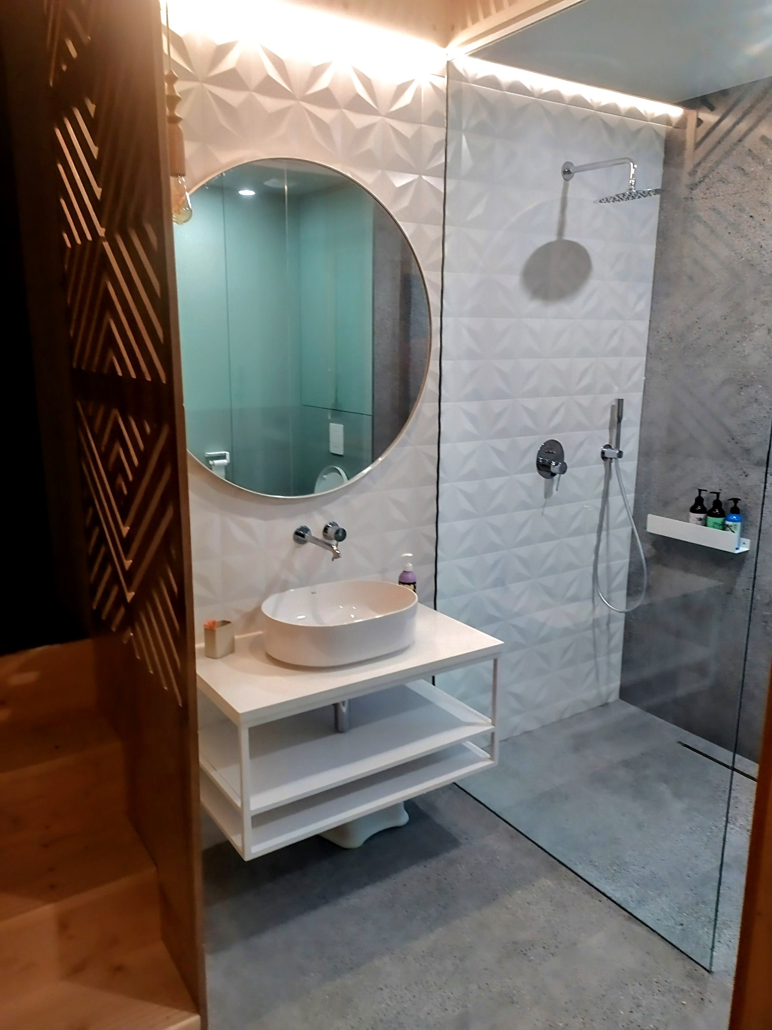 Nowoczesna łazienka z kabiną prysznicową typu walk-in, umywalką na szafce i okrągłym lustrem. Ściany wykończone białymi płytkami 3D i szarym betonem.