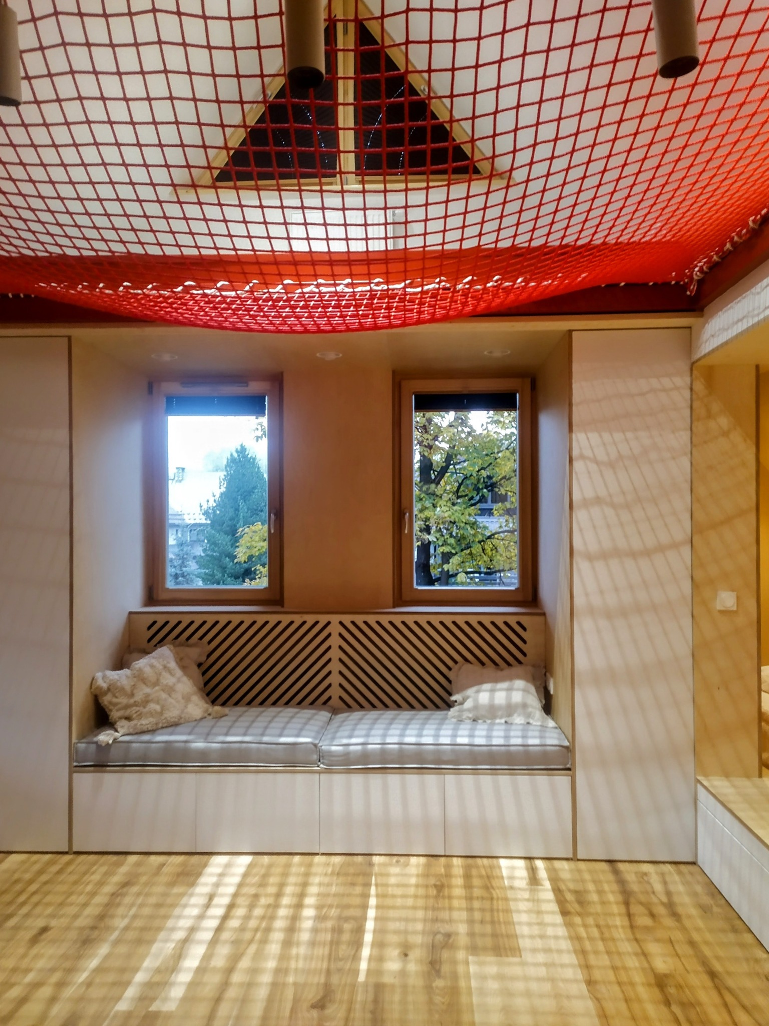 Jasne wnętrze pokoju z drewnianą podłogą, wnęką okienną z siedziskiem i czerwoną siatką pod sufitem. Widok na drzewa za oknami. Nietypowe wykończenie.