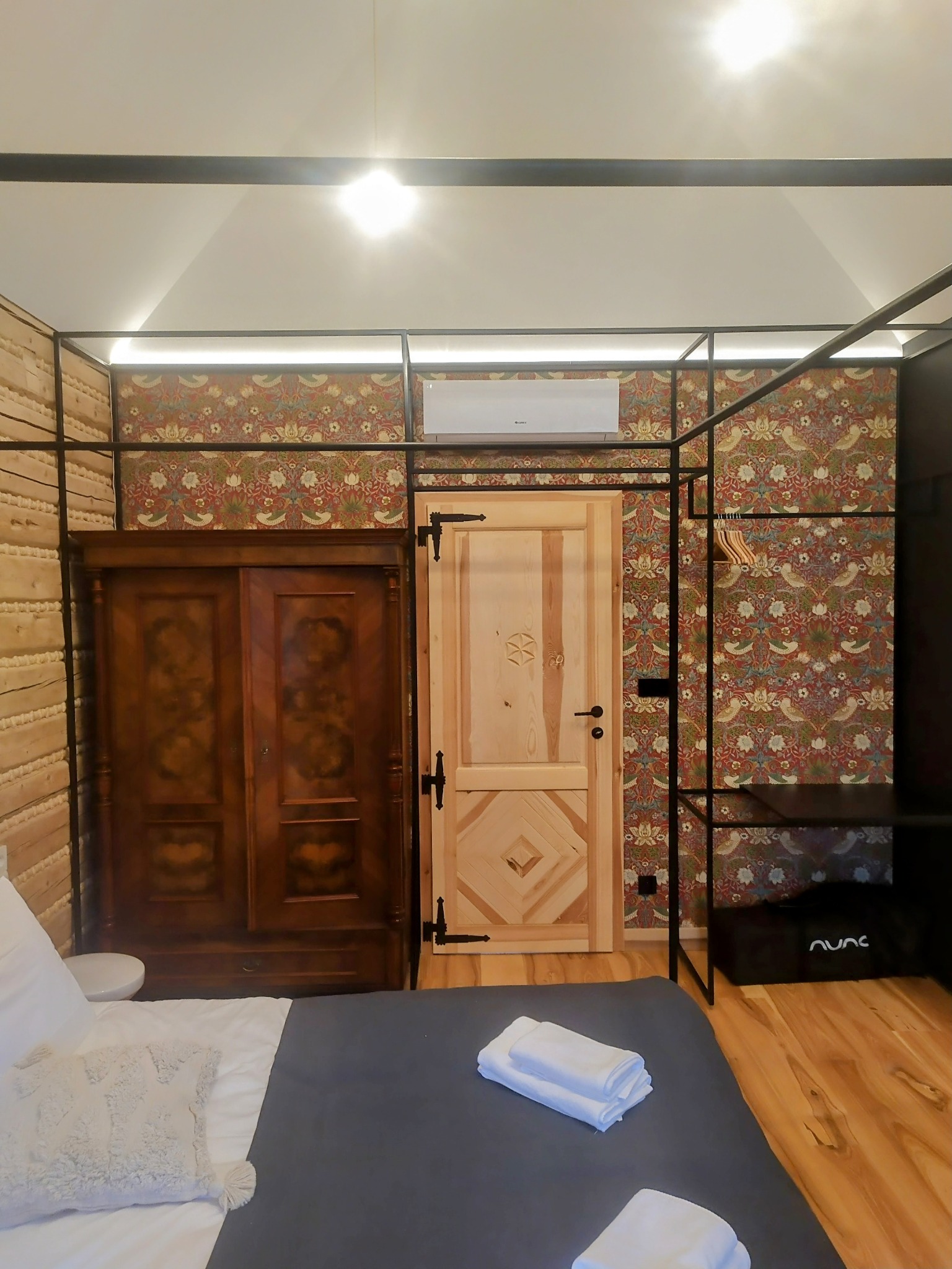 Ukończony pokój gościnny z drewnianą boazerią, wzorzystą tapetą, drewnianymi drzwiami i szafą. Widoczny fragment łóżka. Całość w stylu rustykalnym.