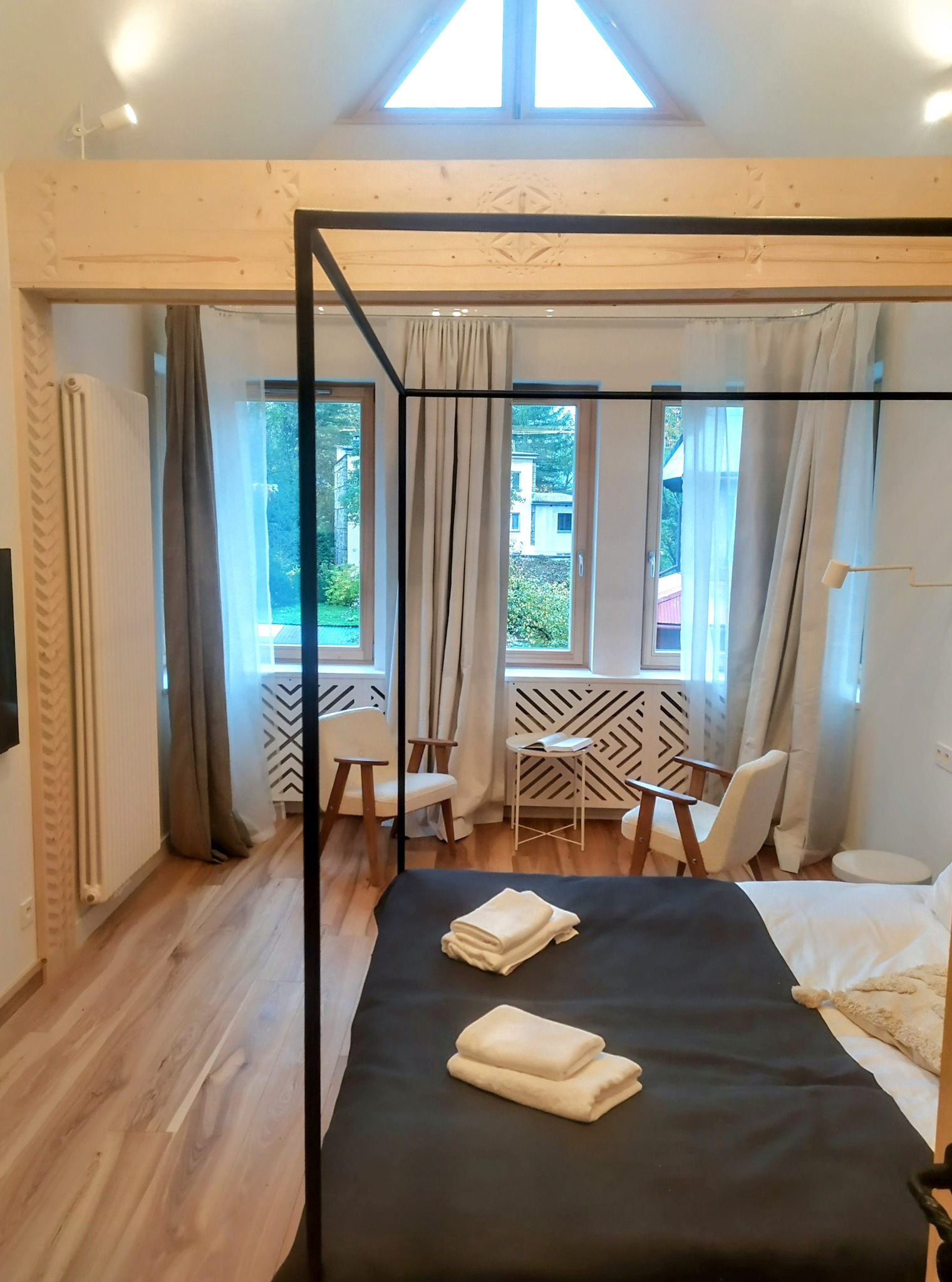 Jasne wnętrze sypialni z drewnianą podłogą, łóżkiem z baldachimem i oknami z widokiem na zieleń. Dekoracyjne elementy drewniane nadają unikalny charakter.