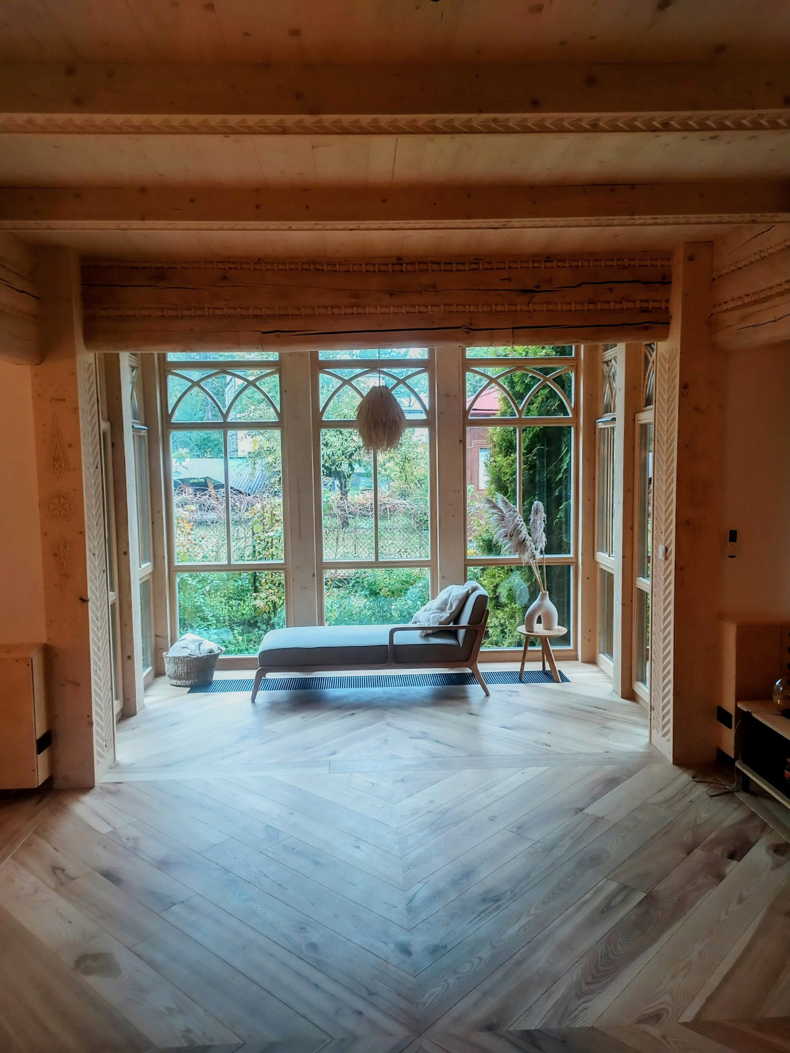 Jasne wnętrze z drewnianym wykończeniem, parkietem w jodełkę i dużym oknem z widokiem na zieleń. W pokoju leżanka i dekoracje w stylu skandynawskim.