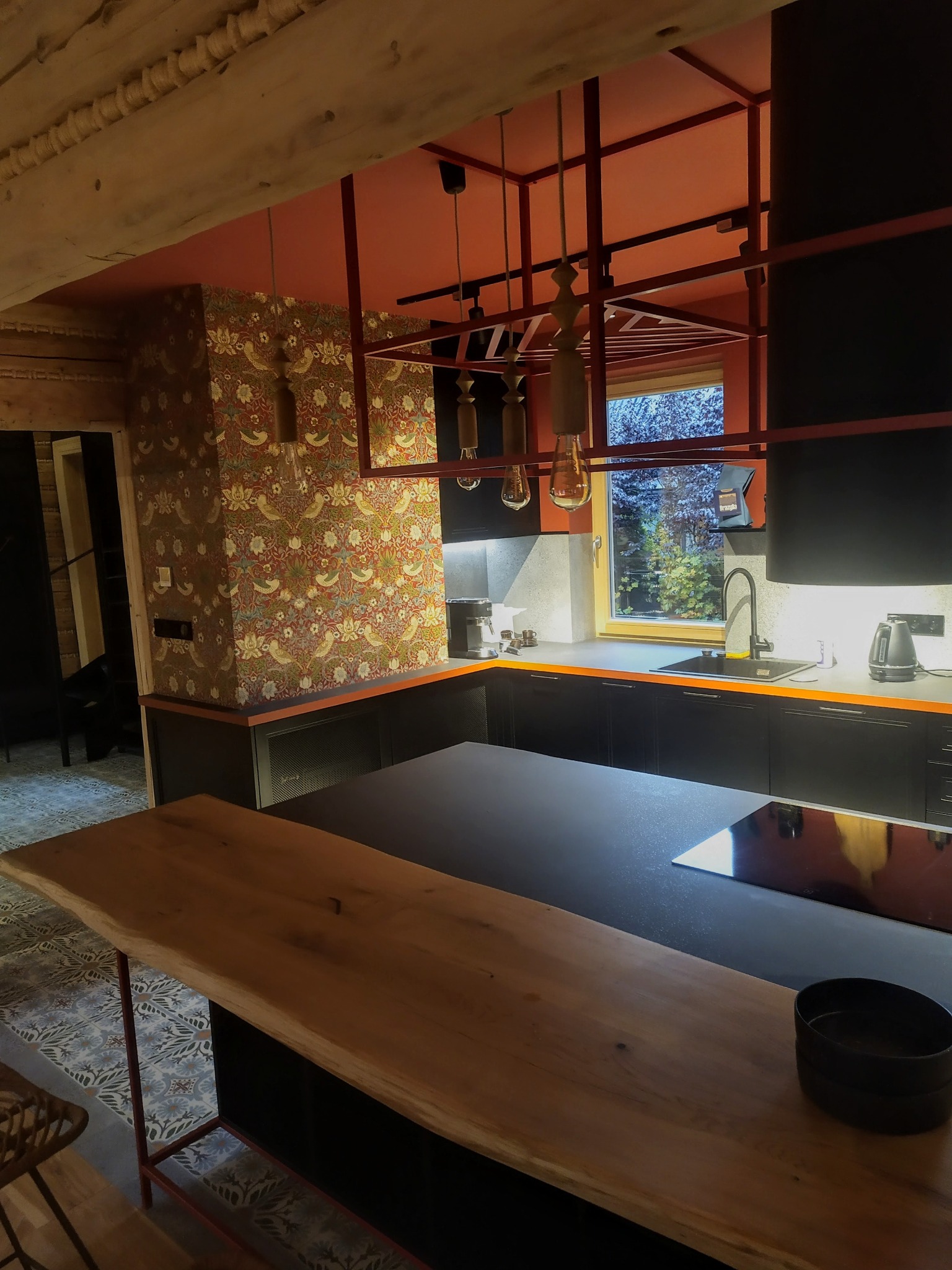 Nowoczesna kuchnia w stylu rustykalnym: czarne szafki, drewniany blat, wzorzysta tapeta i pomarańczowe akcenty. Widoczne oświetlenie i okno z widokiem na zewnątrz.