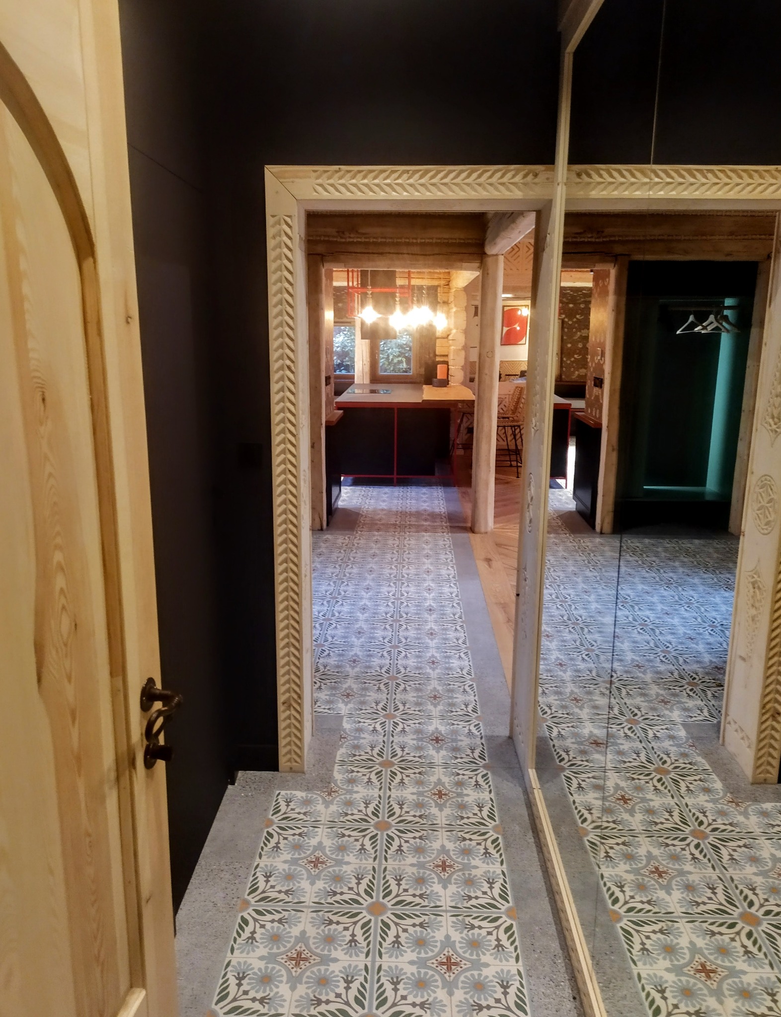 Otwarta drewniana rama drzwi z dekoracyjnym wzorem,  ukazująca wzorzystą podłogę z płytek w głębi wnętrza. Lustro po prawej stronie odbija widok.