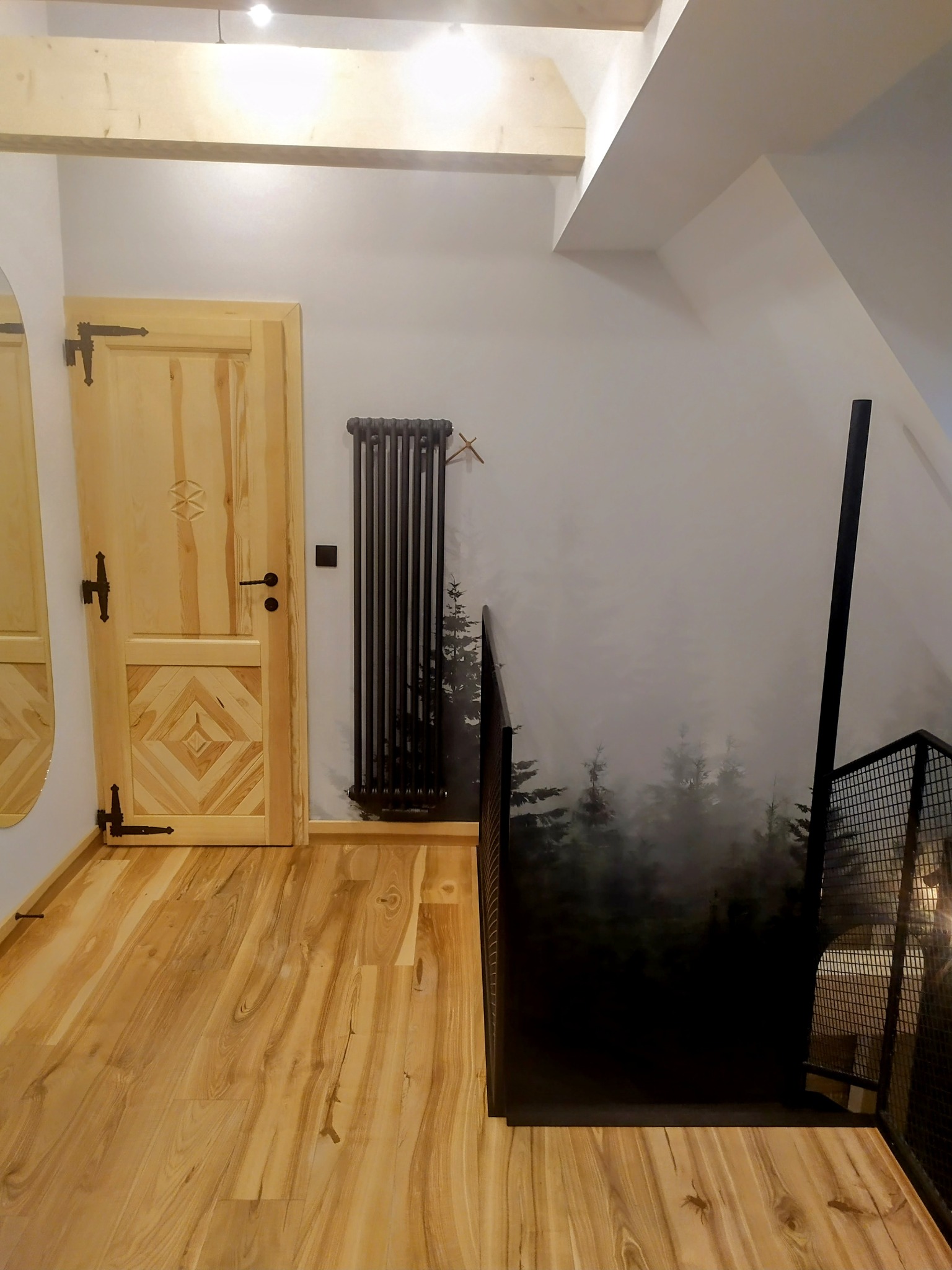 Jasna drewniana podłoga, drzwi z ozdobnymi elementami i ciemny grzejnik na tle ściany z motywem lasu. Wykończenie wnętrza w stylu górskim.