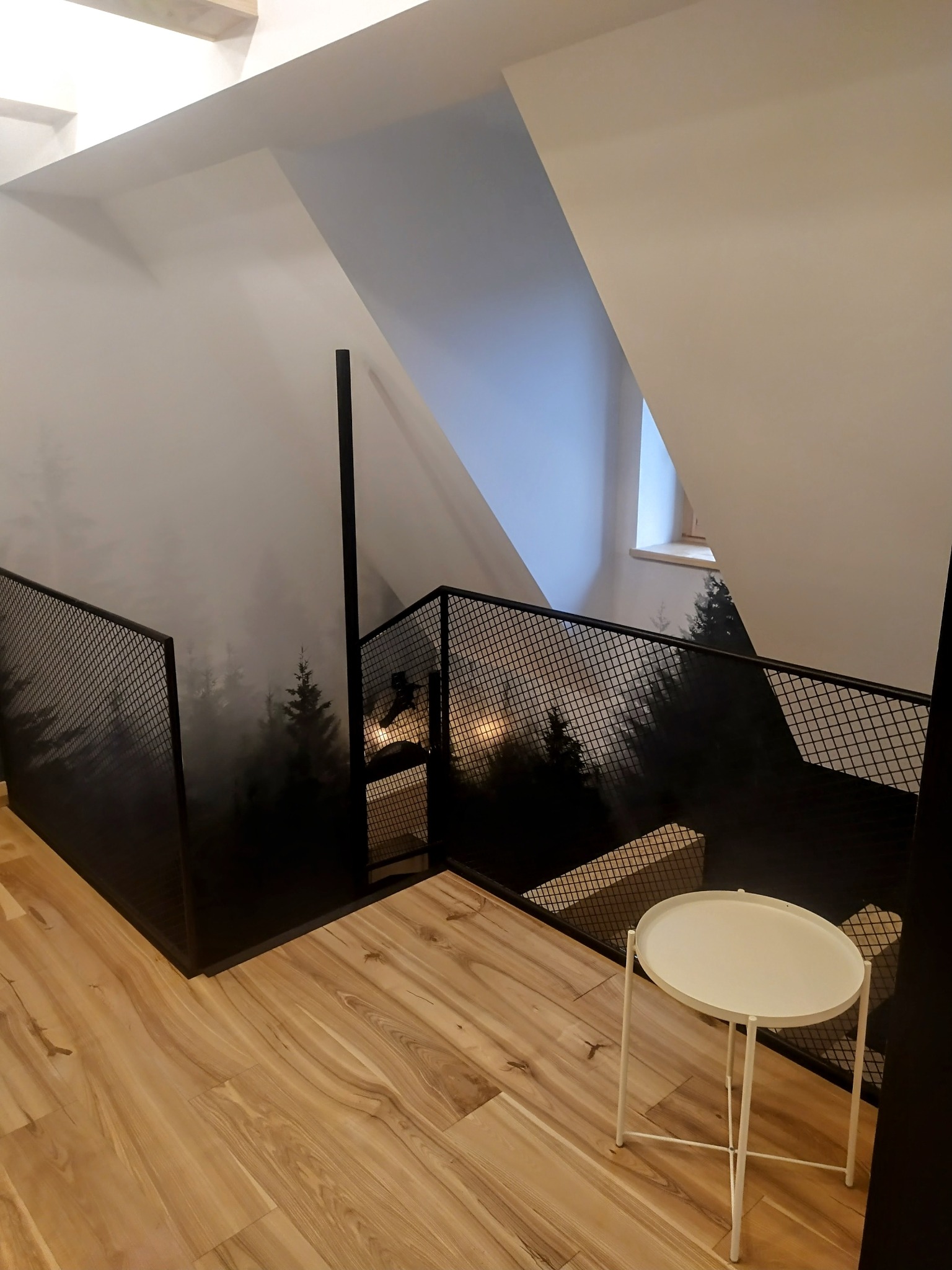 Klatka schodowa w domu z drewnianą podłogą i czarną, metalową balustradą z motywem lasu. Białe ściany i stolik kawowy. Widok na okno.