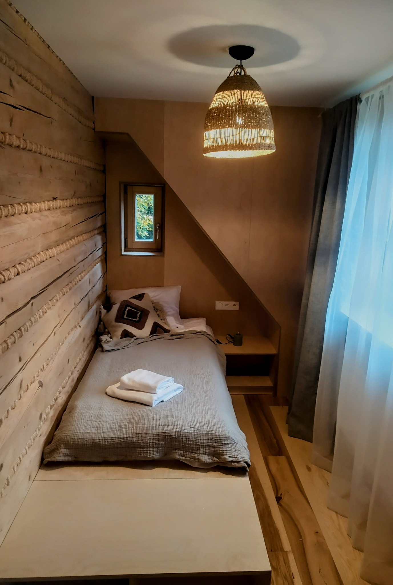 Przytulna sypialnia w stylu góralskim: drewniane ściany, okno, łóżko z lnianą pościelą i lampa z wikliny. Subtelne oświetlenie i naturalne materiały tworzą ciepły klimat.