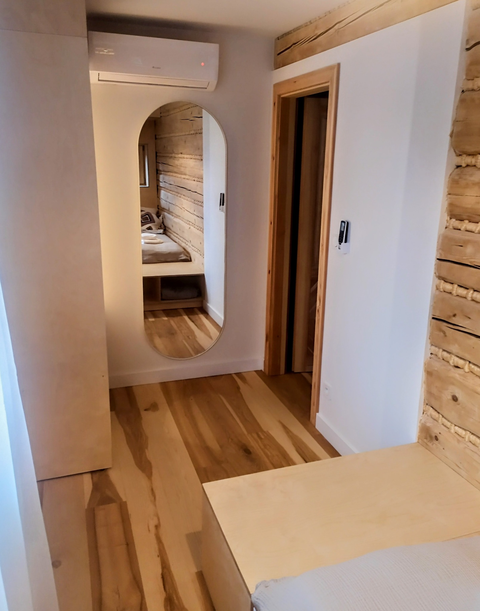 Wnętrze z drewnianą ścianą odbijającą się w owalnym lustrze, widoczne drewniane podłogi i drzwi. Klimatyzator na ścianie. Wnętrze w stylu skandynawskim.