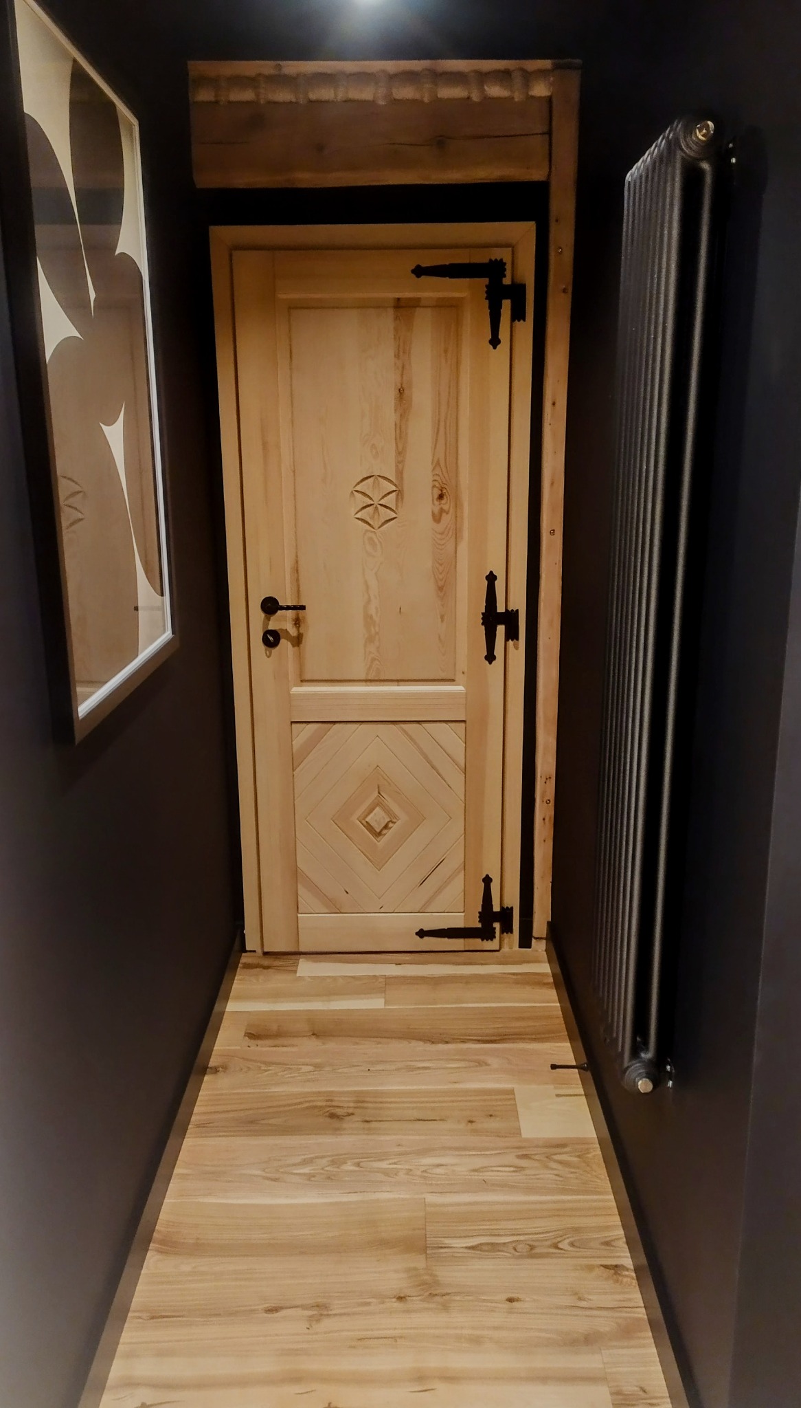 Jasne drewniane drzwi z dekoracyjnymi okuciami w ciemnym korytarzu z drewnianą podłogą. Na ścianie obraz w ramie i grzejnik żeberkowy.