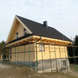 Dom w trakcie budowy, widoczne drewniane ściany, czarny dach i rusztowania, okna zabezpieczone folią, fundament z bloczków betonowych.